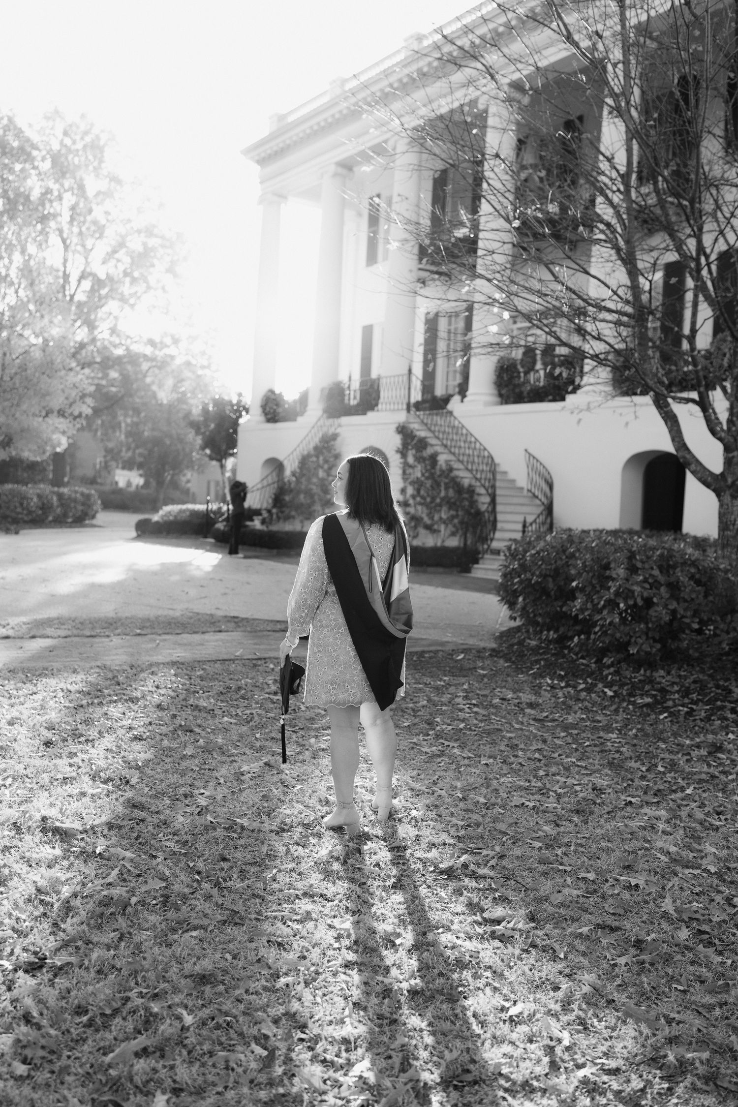 A graduate walks through a scenic autumn campus lawn covered in fallen leaves near white columned buildings.