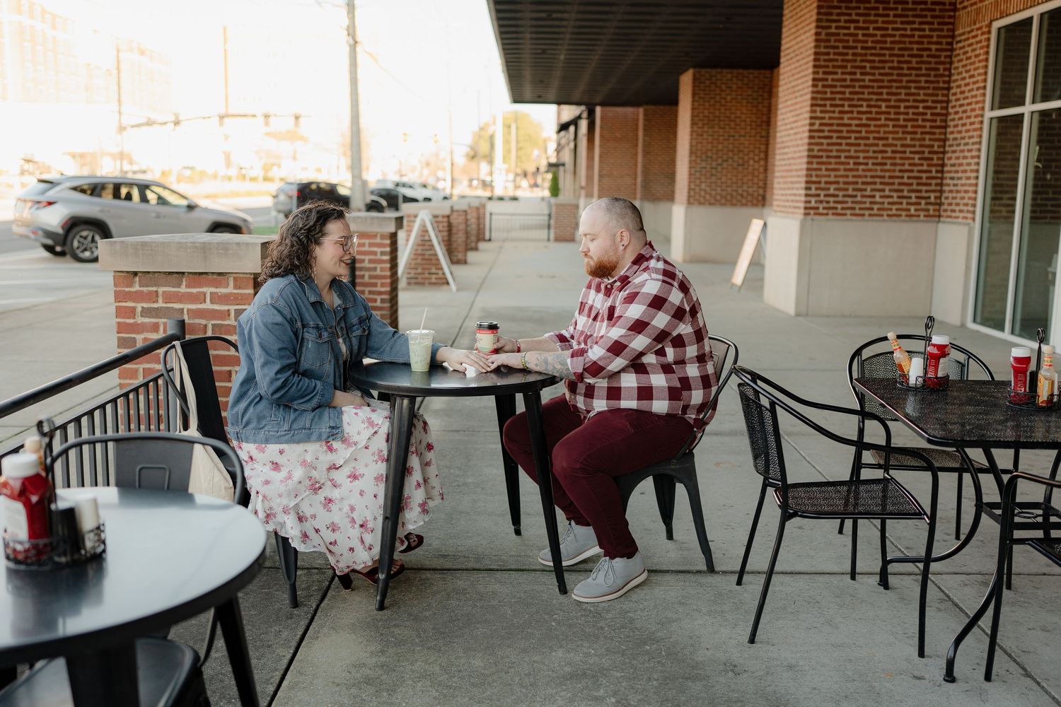 Two people enjoy coffee and conversation at an outdoor cafe patio with metal tables and chairs on a sunny day.