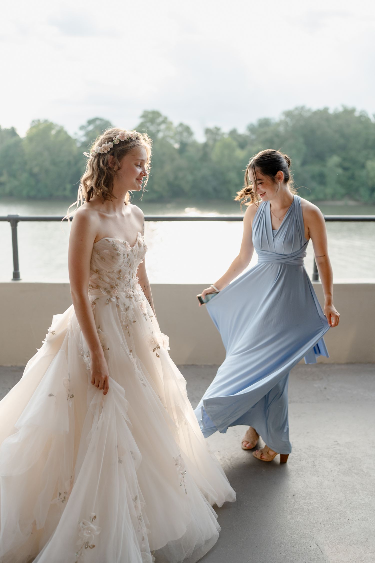Two women in formal dresses, one in a flowing wedding gown and one in light blue, dancing together by a lake.