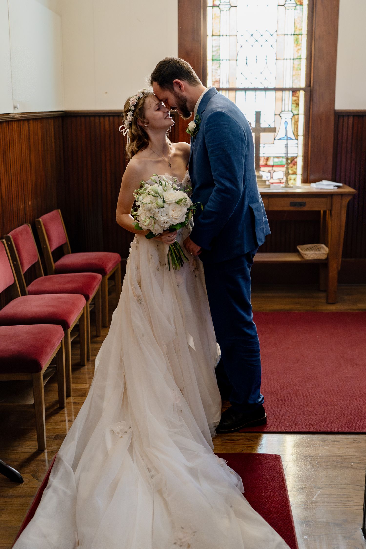 A couple shares their first look moment in a church with stained glass windows and red carpeted floors.