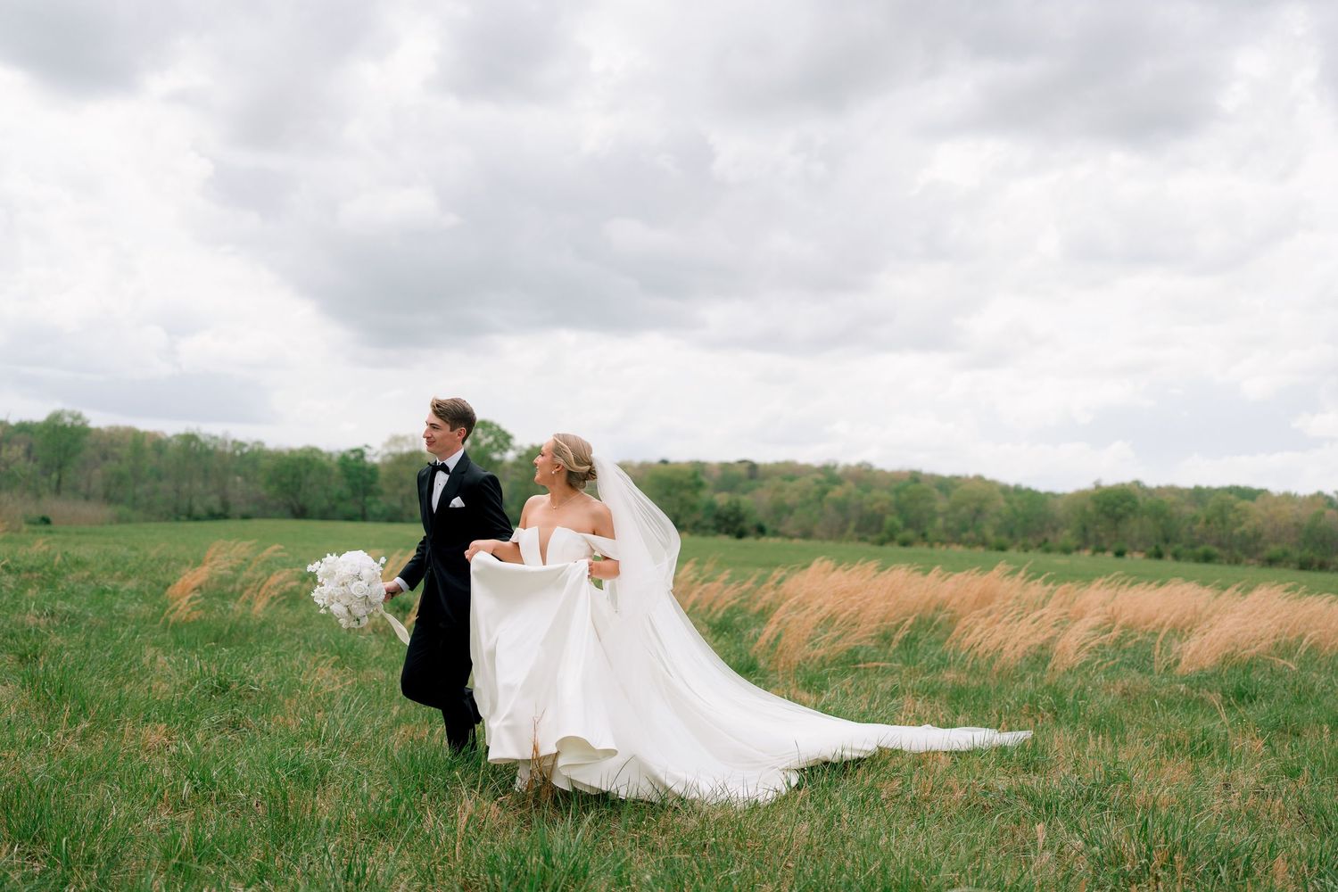 Bride and groom walk through lush green field on windy wedding day, dress flowing, bouquet in hand.