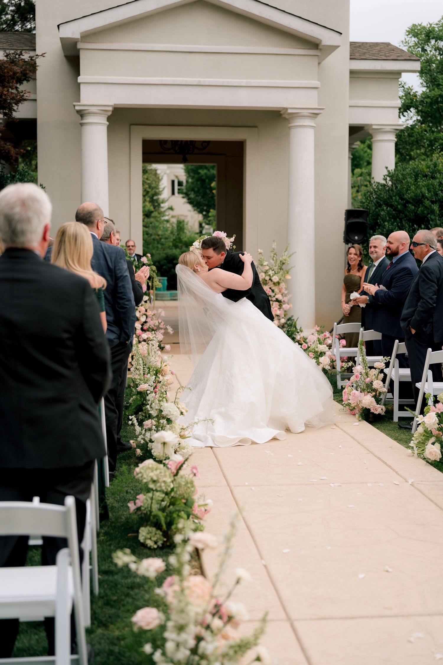 Bride and groom share first kiss walking down floral-lined aisle at elegant outdoor wedding ceremony.