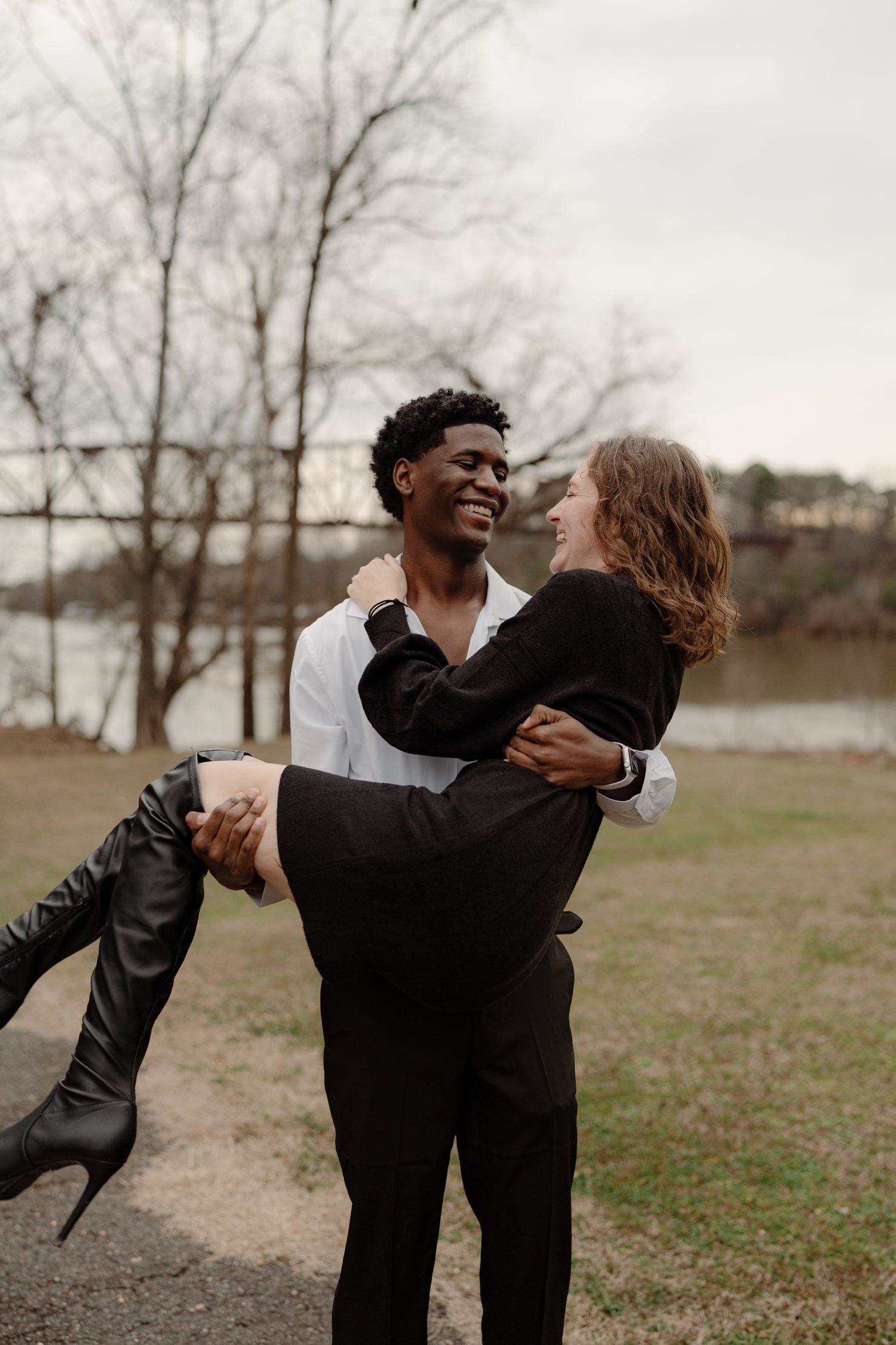 A joyful interracial couple embraces outdoors, the man lifting the woman near a serene lake.
