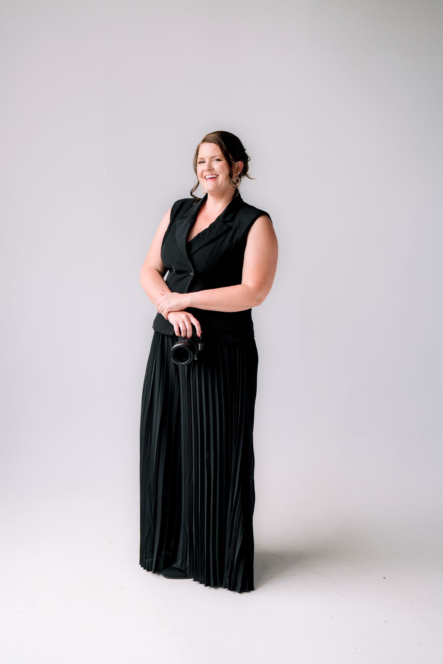 Woman in elegant black sleeveless pleated gown holding a camera, smiling confidently in a studio setting.