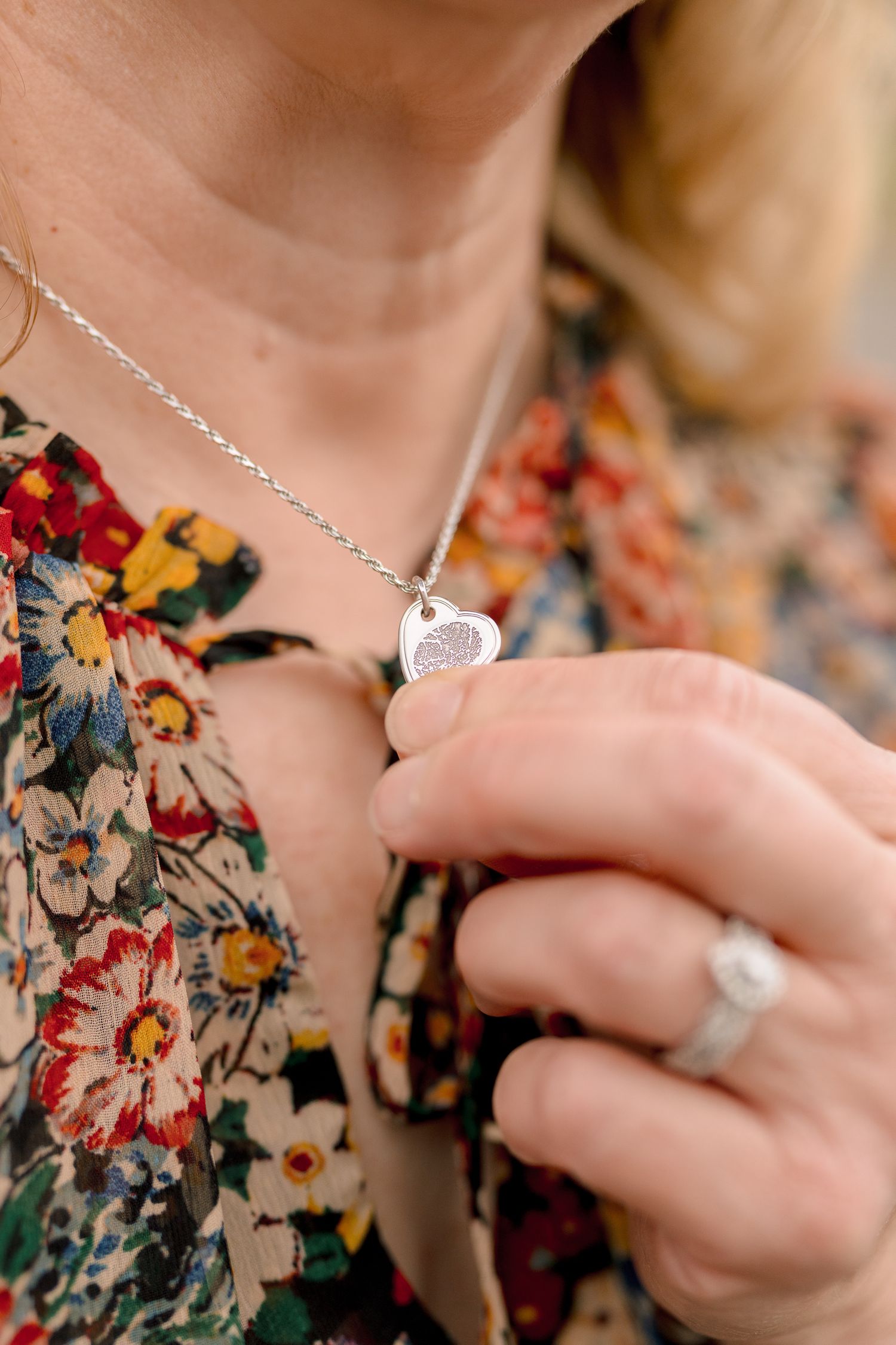 Close-up of woman displaying a silver heart fingerprint pendant necklace while wearing a floral dress.