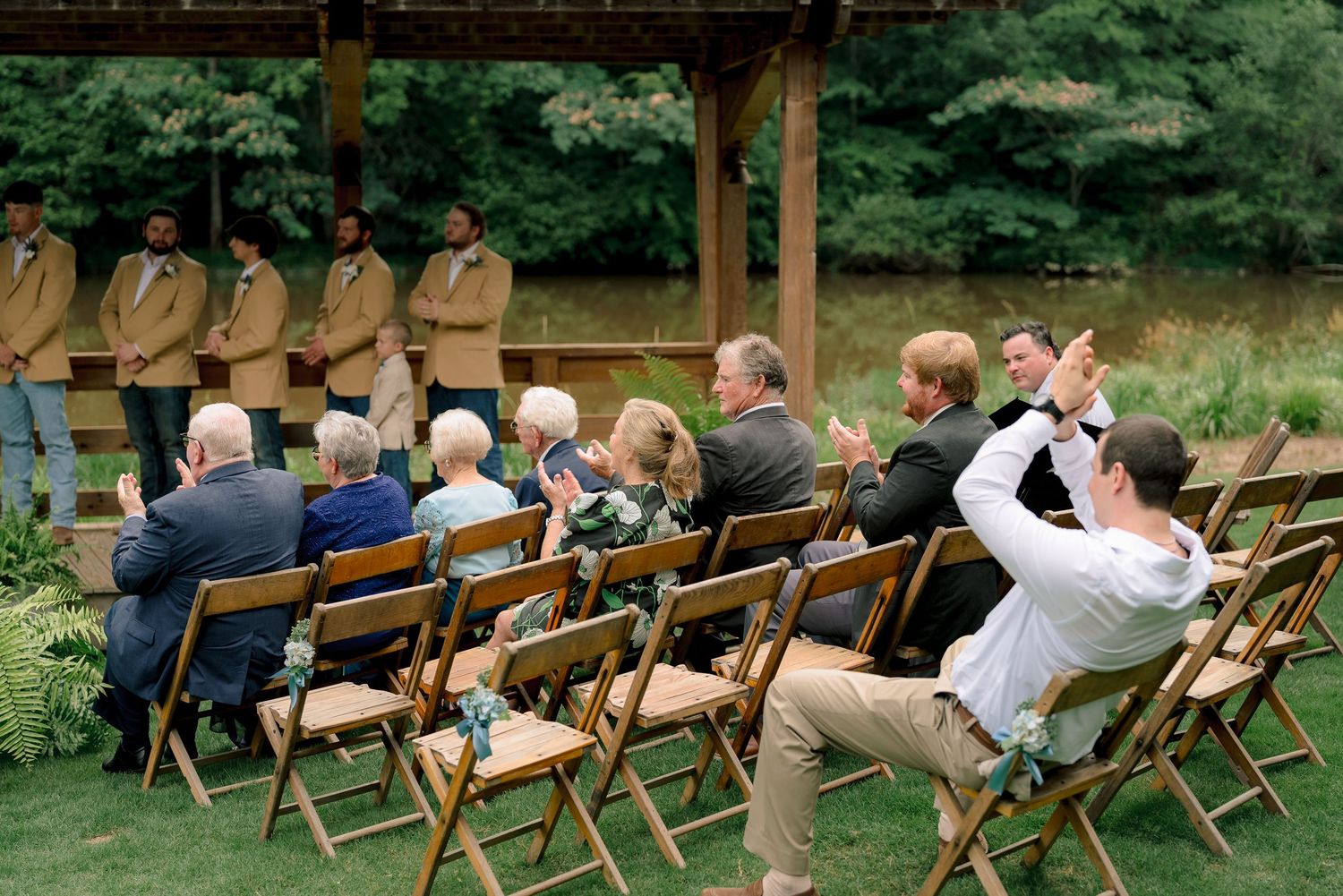 Wedding guests seated in wooden folding chairs applaud during outdoor ceremony near rustic wooden pavilion.