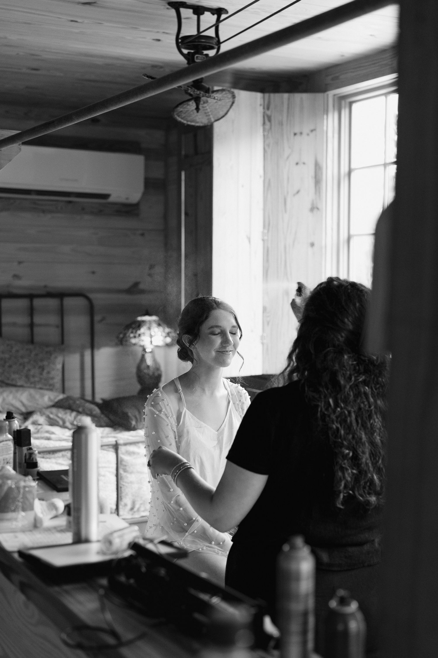Black and white photo of bride getting makeup done by artist in rustic wood-paneled room before wedding.