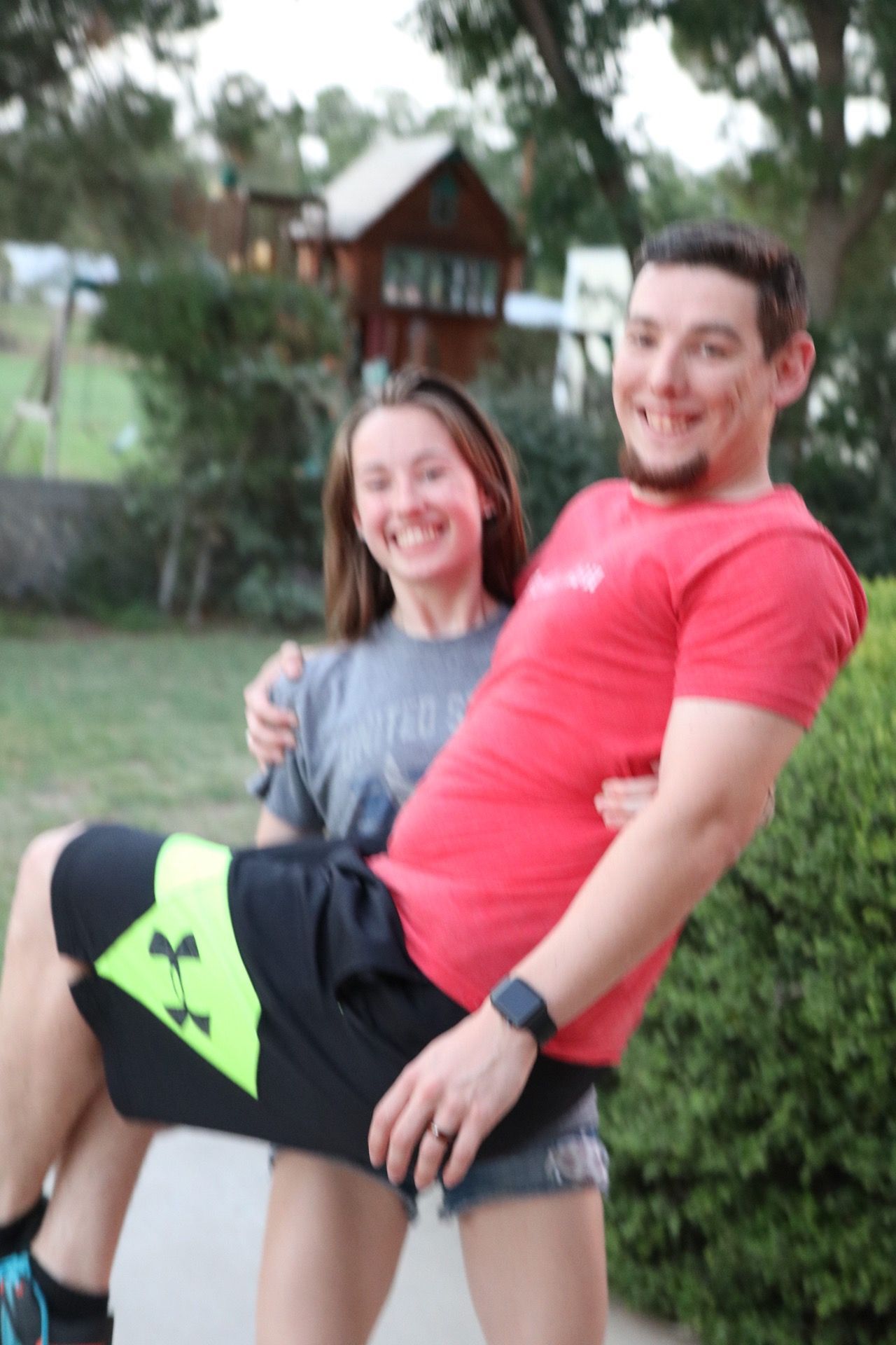 Two people pose together outdoors with a red cabin visible in the background.