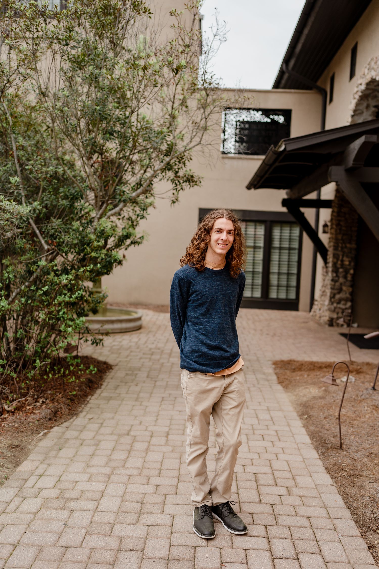 Person in casual attire standing on brick walkway near flowering garden and beige building.