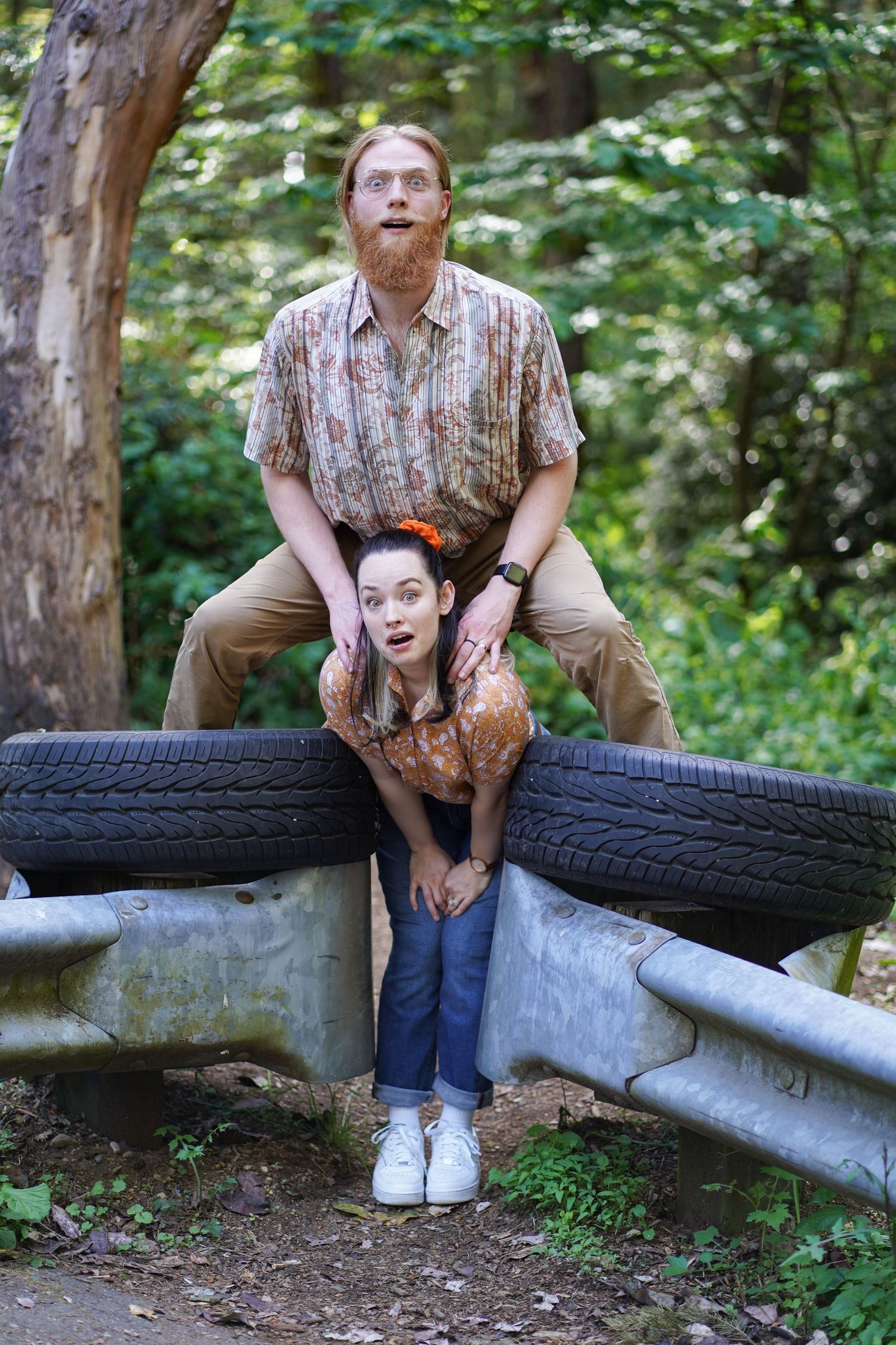 A playful outdoor portrait with two people posing by stacked tires and guardrails in a wooded area.