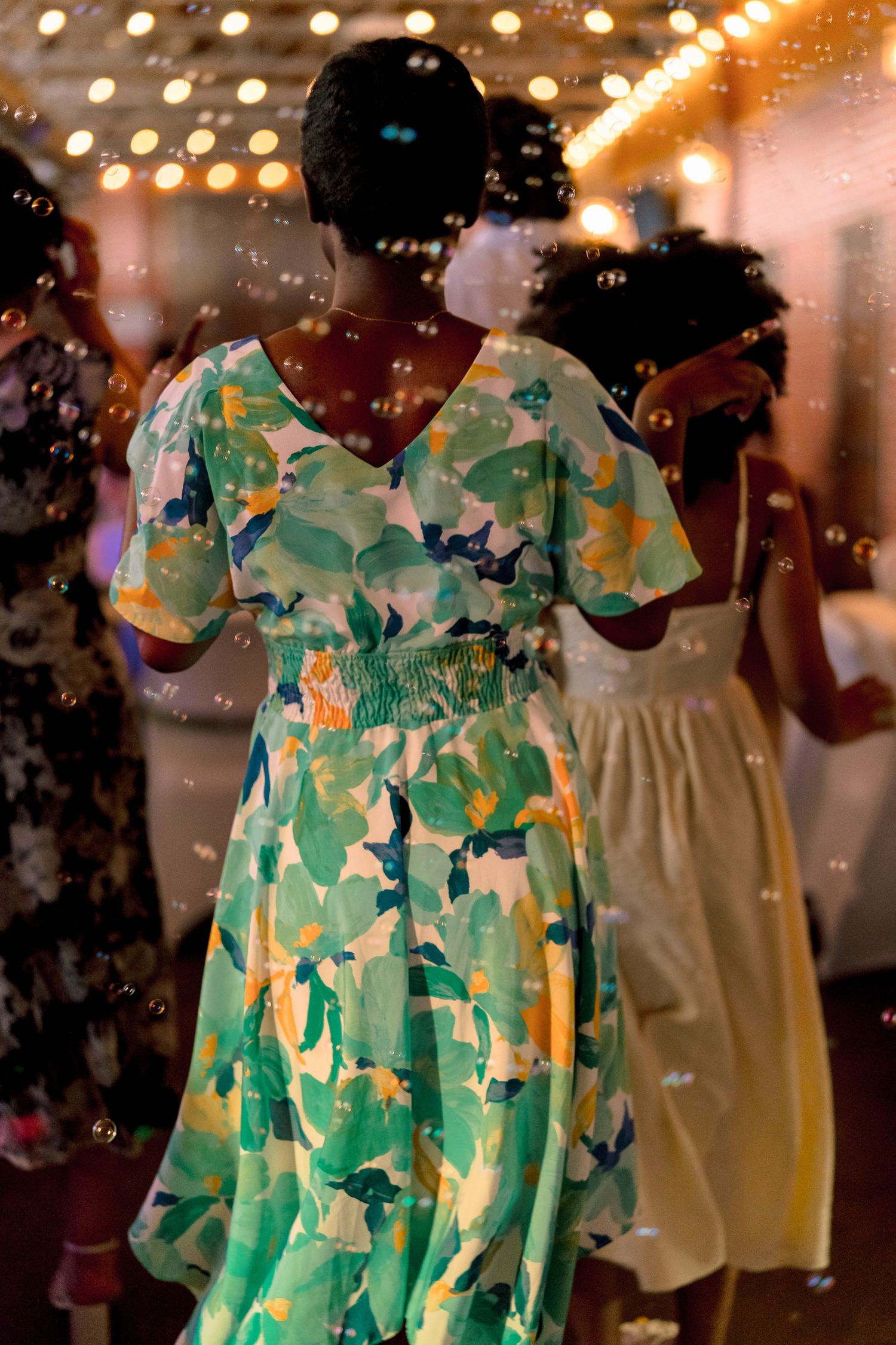 Guest in colorful floral green dress dances surrounded by bubbles at wedding reception.