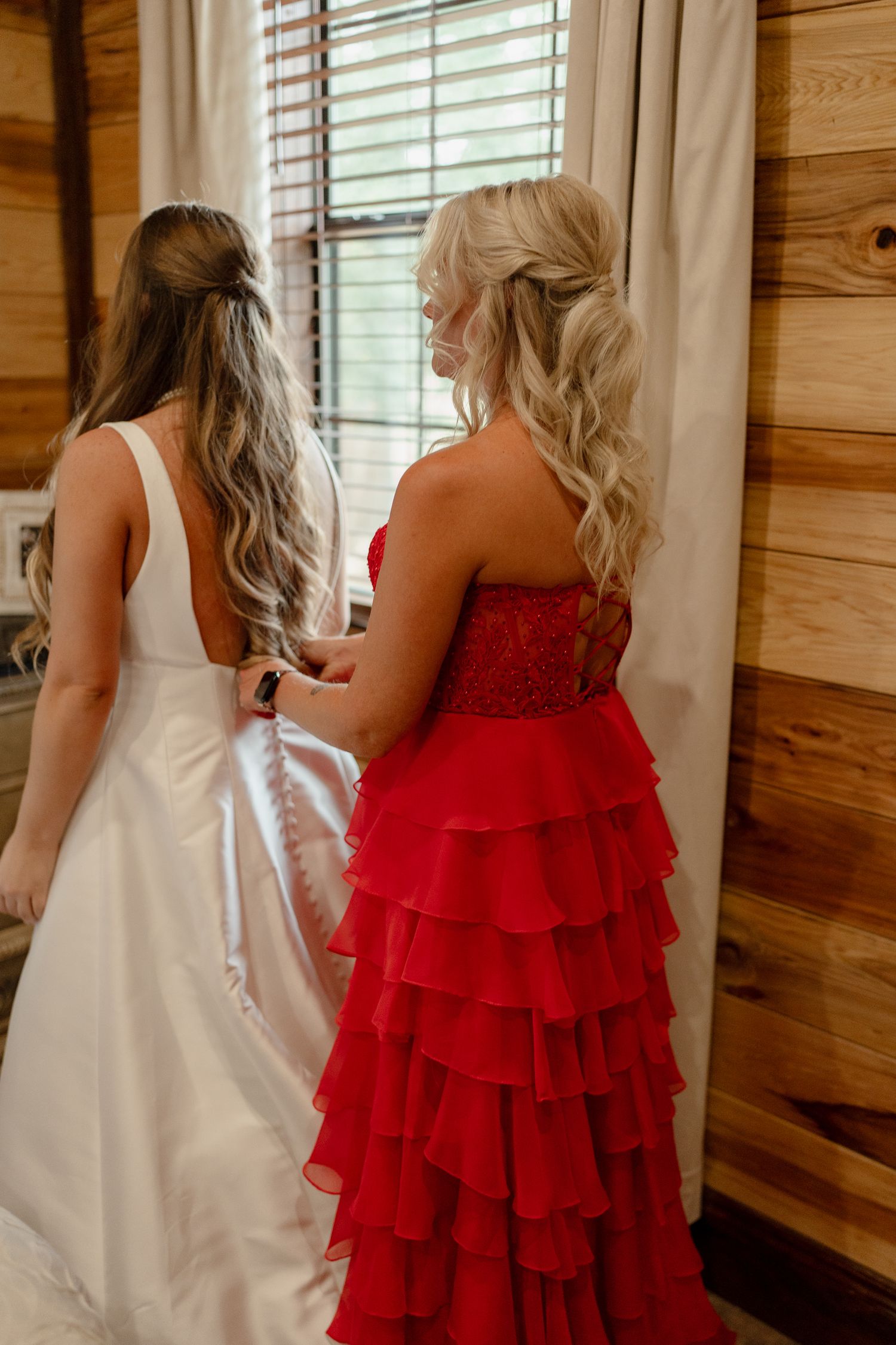 A bride in white and bridesmaid in red ruffled dress stand together in a rustic wooden interior.