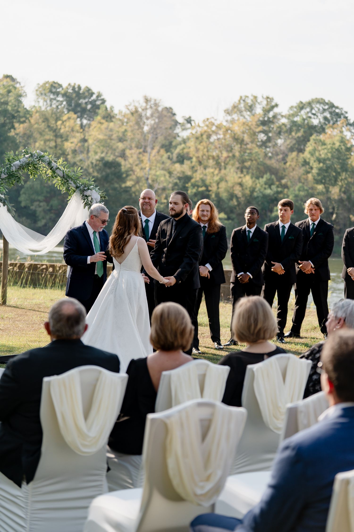 Wedding party stands in a line outdoors while guests watch from white chairs during an autumn ceremony.
