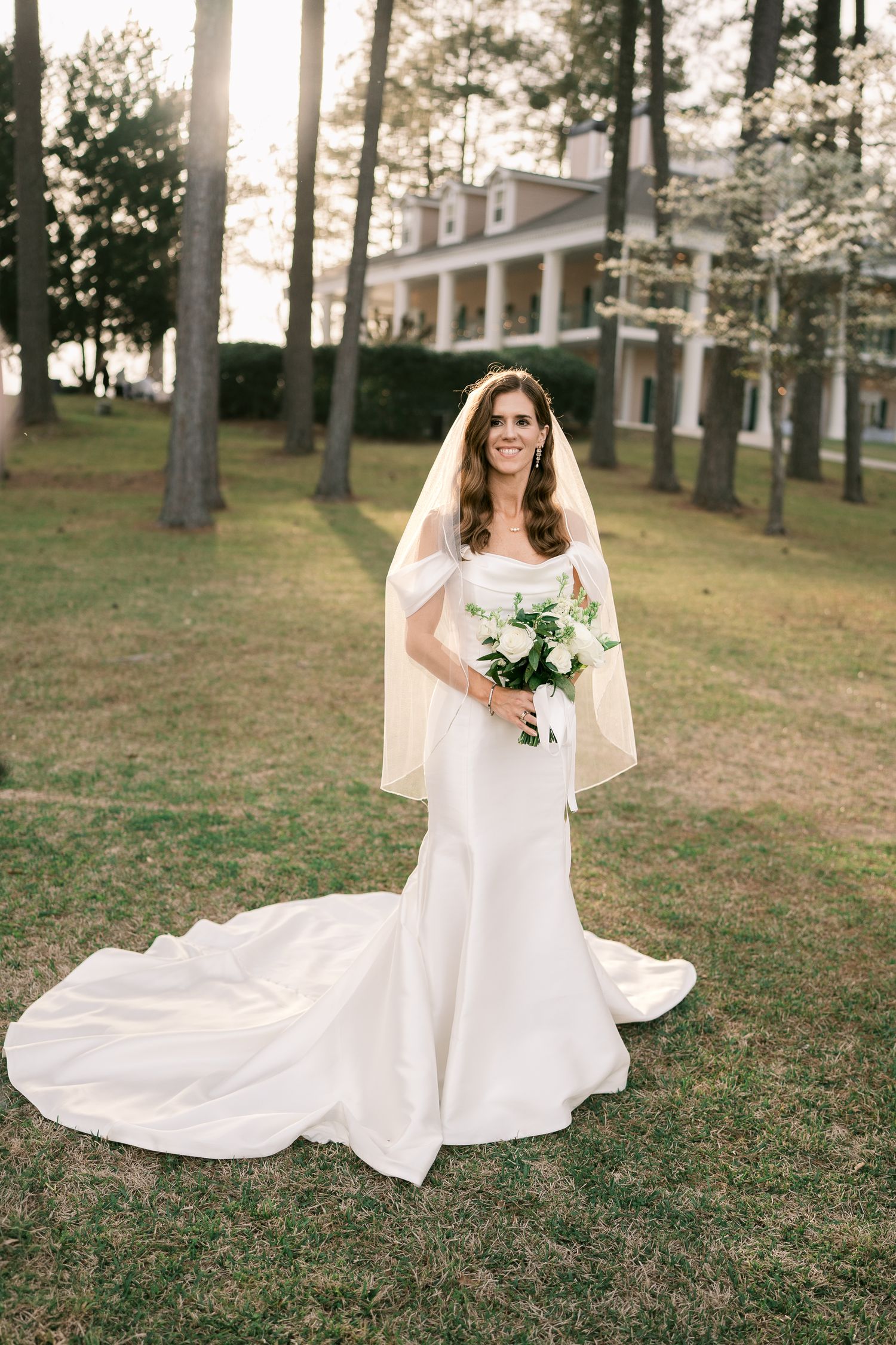 Bride in elegant white dress stands with flowing train against backdrop of historic mansion.