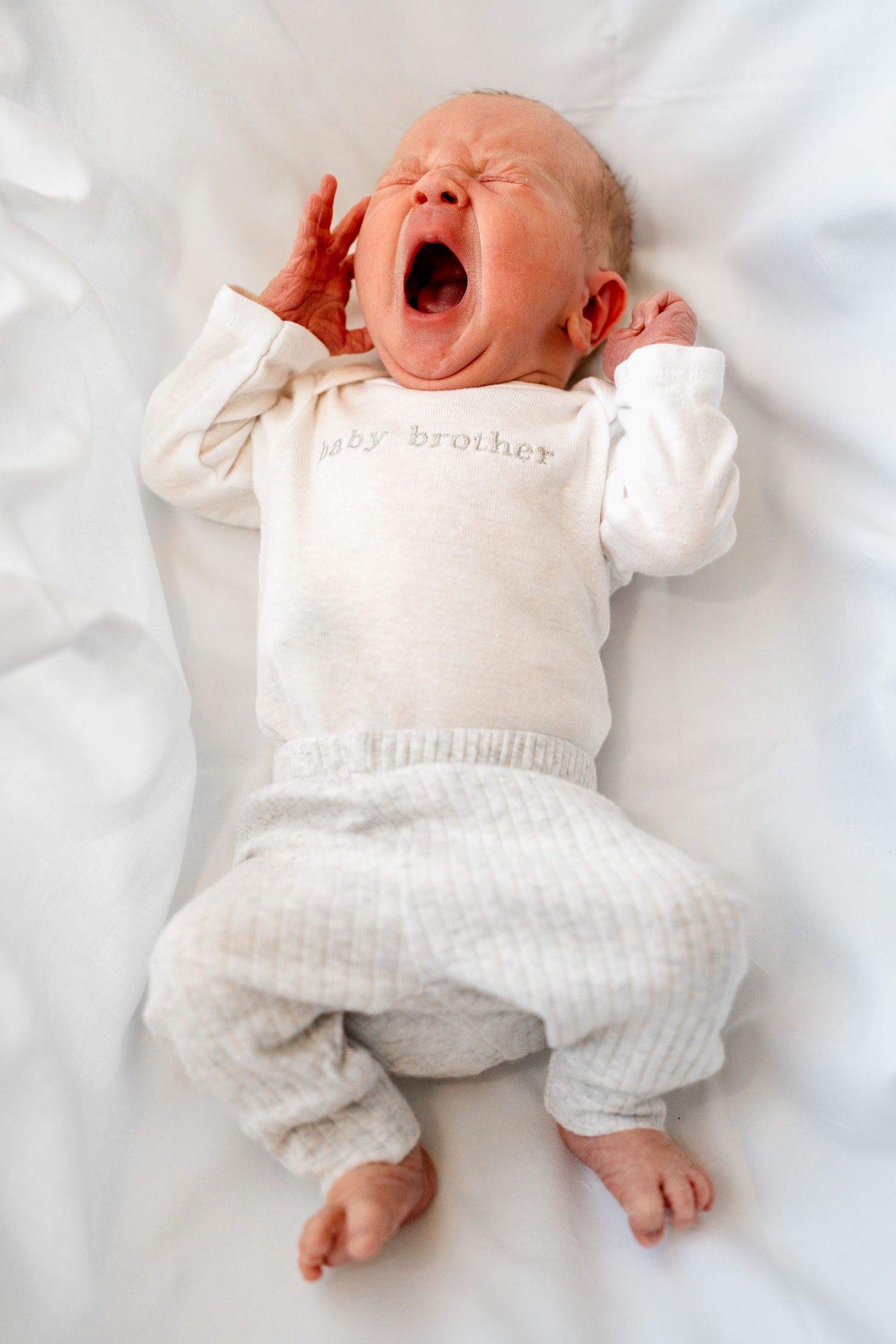 Baby in white outfit yawning while lying on white bed sheets.