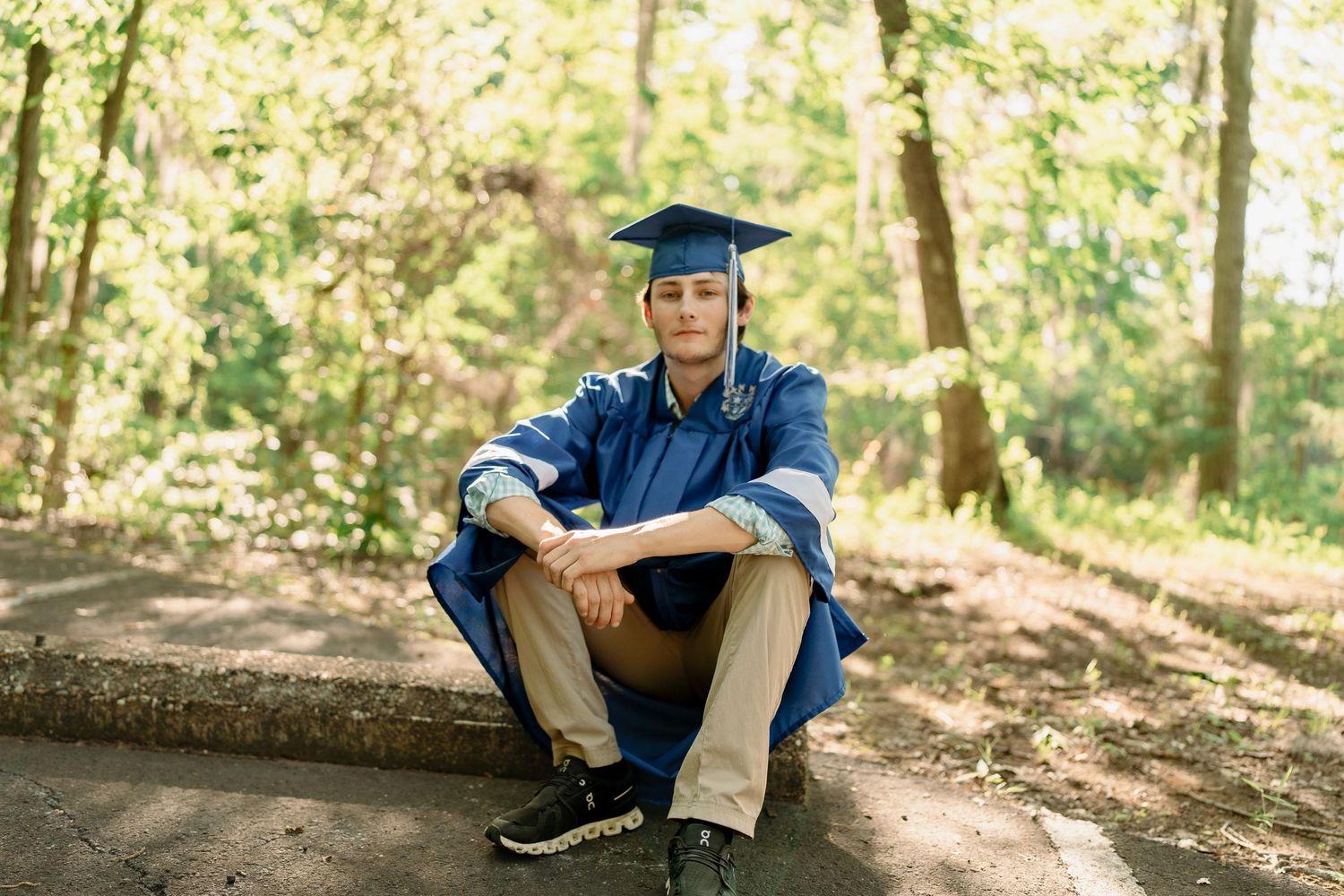 A graduate in blue academic regalia sitting on a path surrounded by trees in natural lighting.