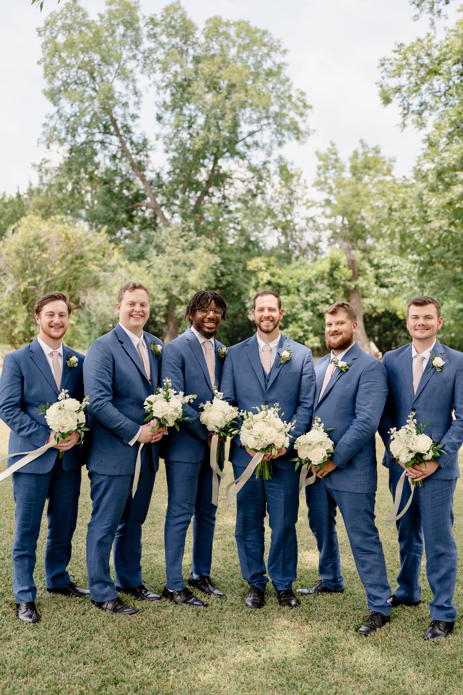 Groomsmen in matching navy blue suits hold white floral bouquets while standing on grass with trees in background.