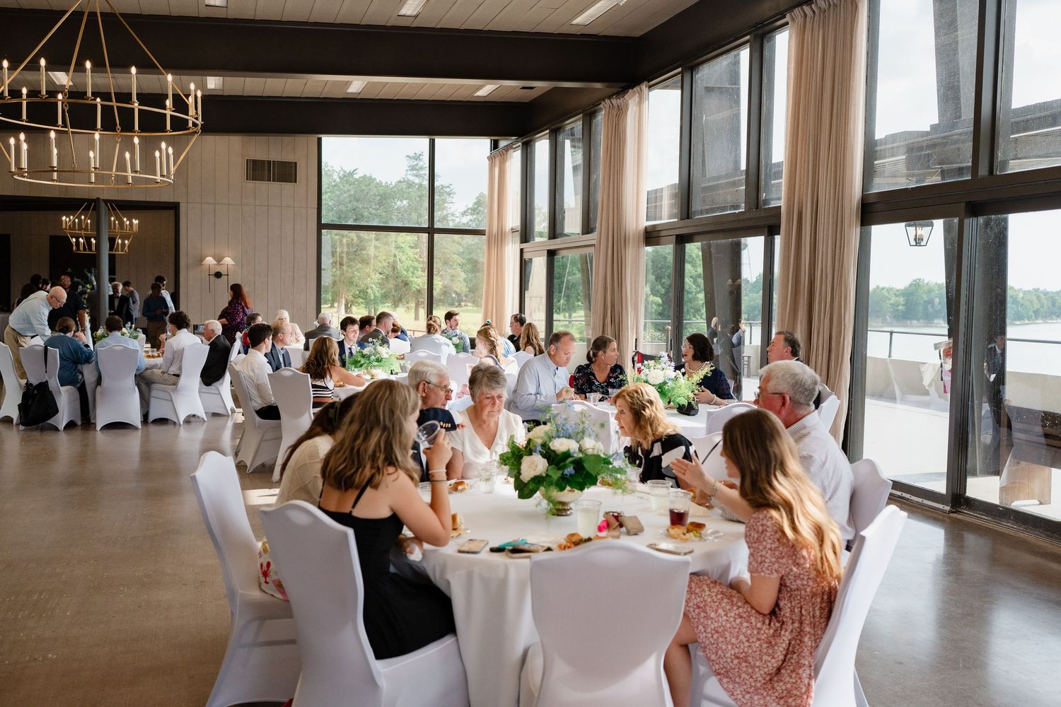 Wedding guests sit at round tables in an elegant reception venue with large windows and chandeliers.