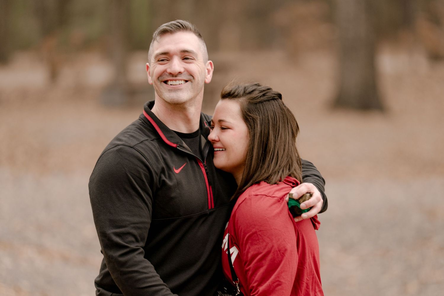 A couple wearing black and red clothing embraces outdoors during an autumn photoshoot.