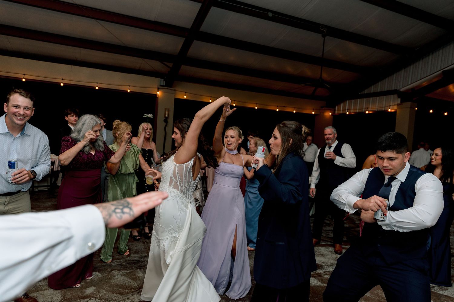 Bride in white gown dancing joyfully with guests at a rustic wedding reception with string lights.