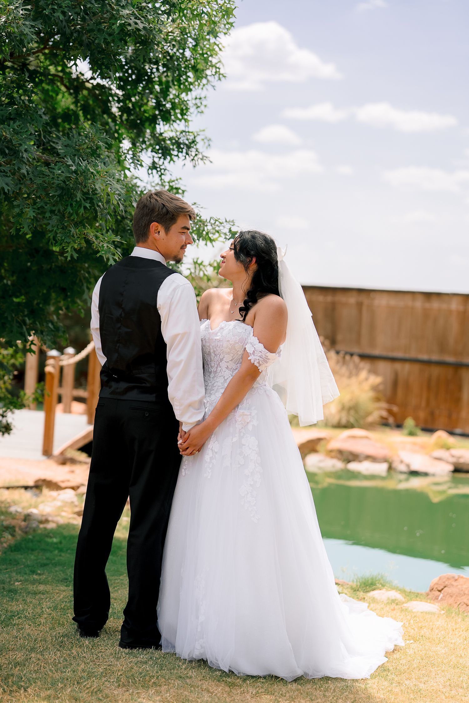 Bride and groom holding hands outdoors, sharing a romantic moment in wedding attire near a pond.