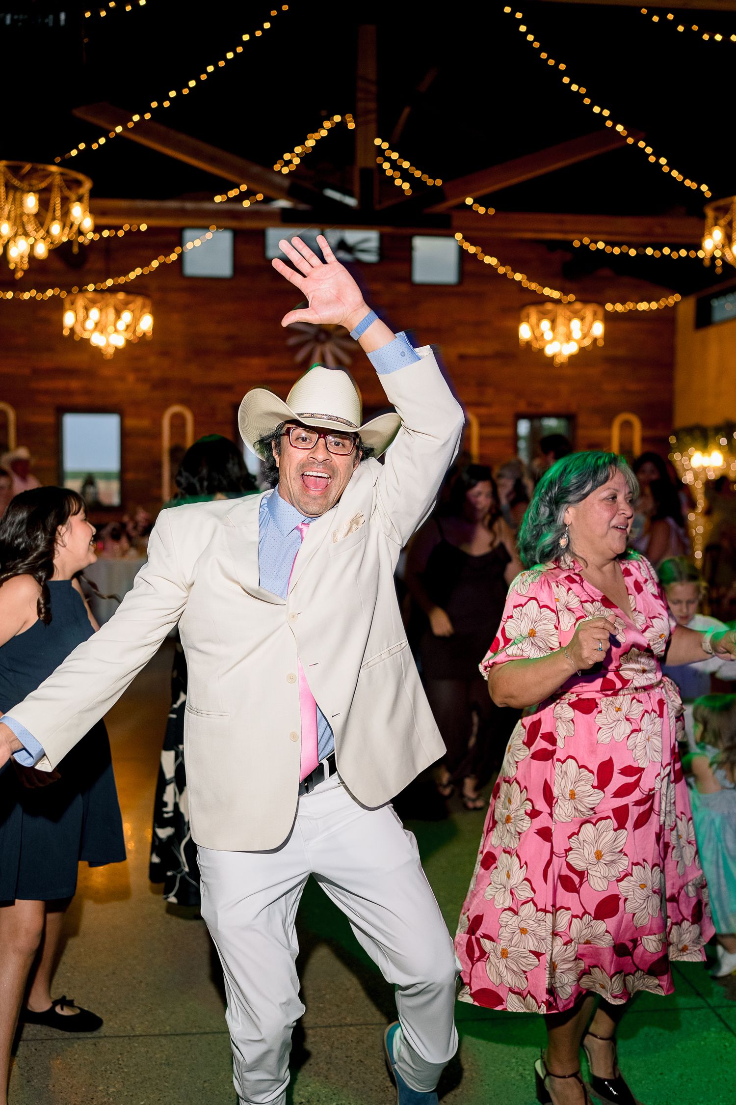 An energetic wedding guest in a white cowboy hat and suit dances enthusiastically on a decorated barn reception dance floor.