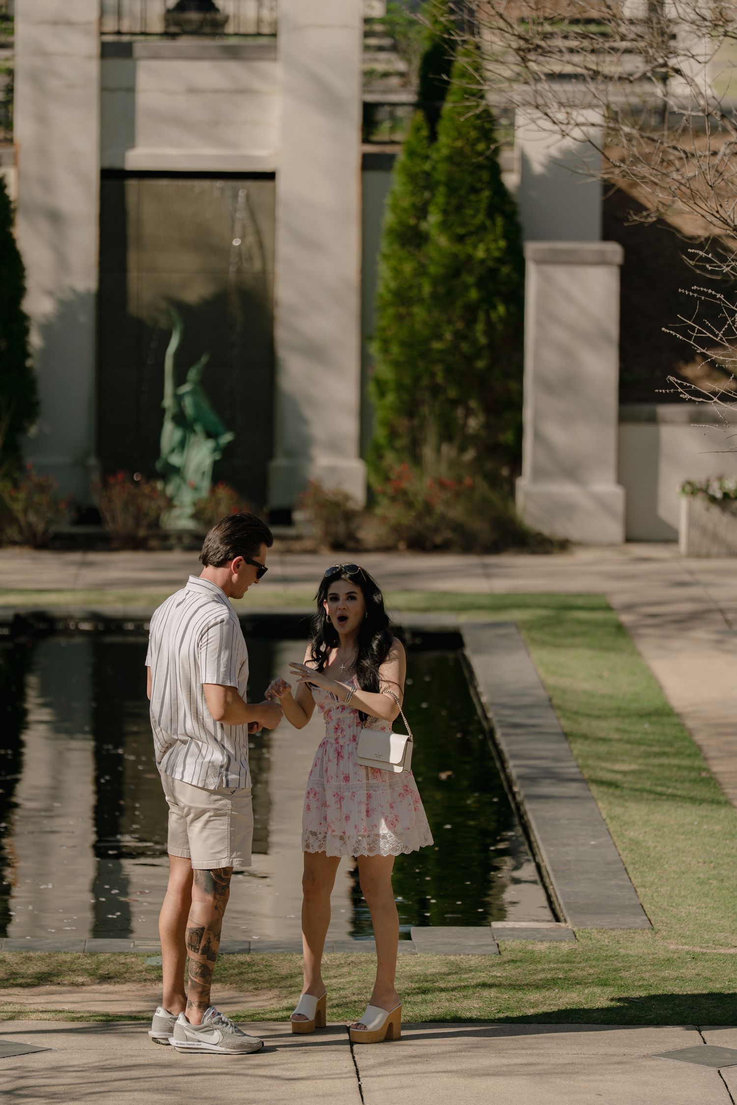 Couple standing by a reflective pool in a formal garden, man in shorts and woman in floral dress on a sunny day.