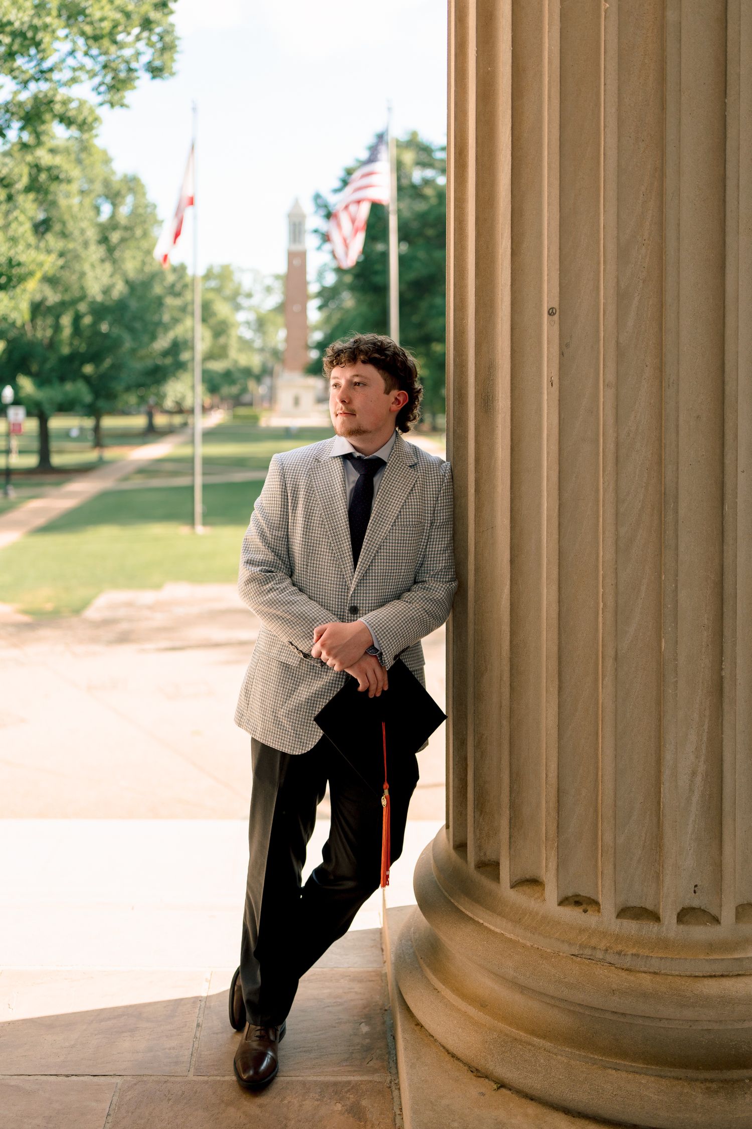 Young man in gray blazer and tie leans against a column holding graduation regalia with American flag behind.
