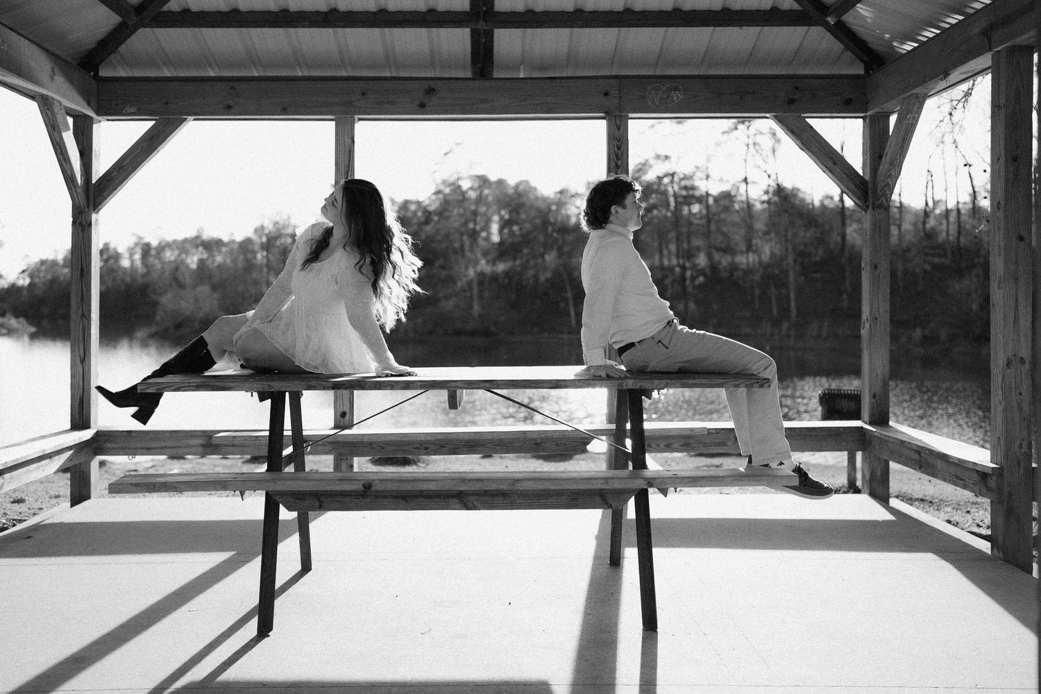 Black and white photo of couple sitting back-to-back on a bench inside a lakeside gazebo.