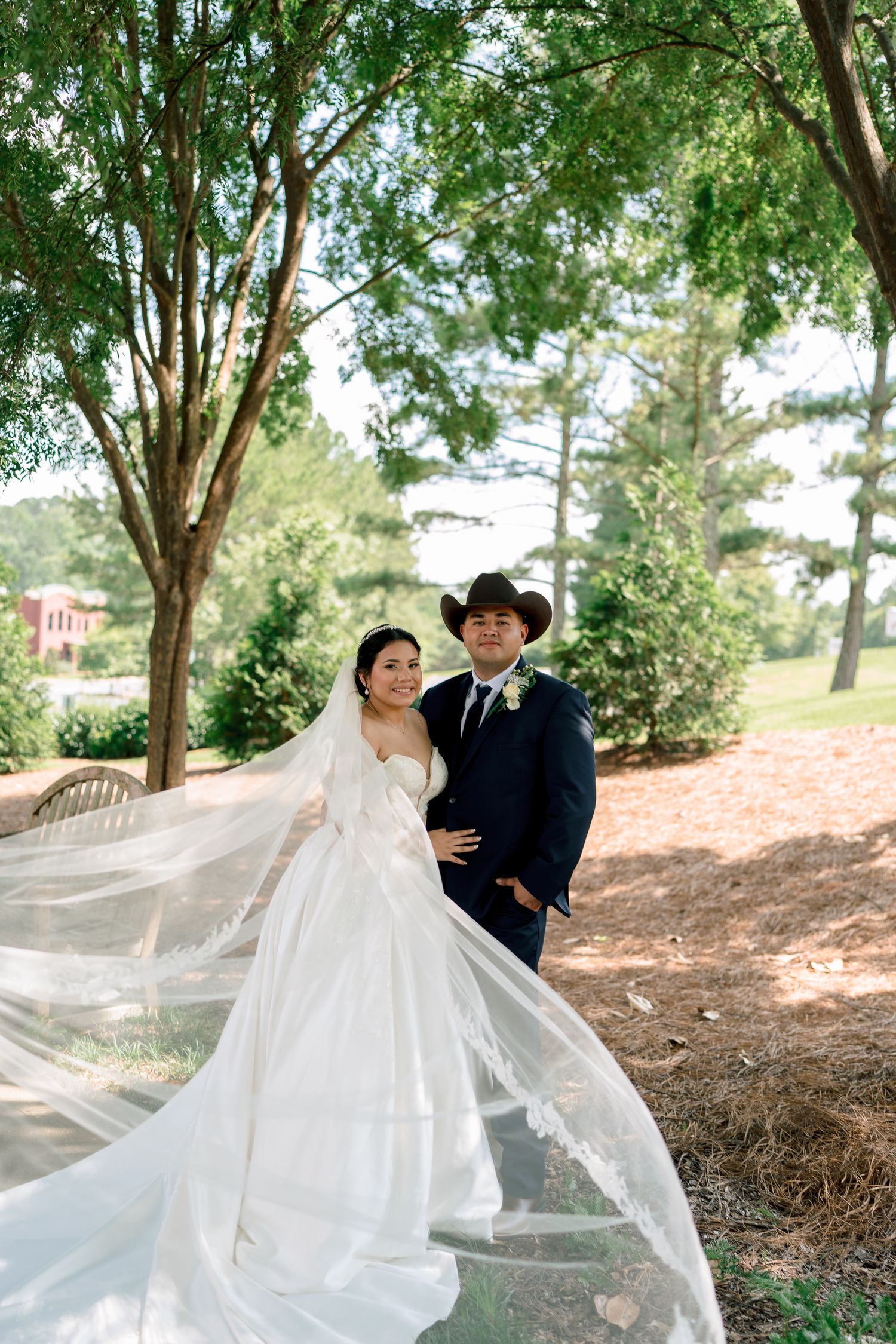 A bride's white veil flows gracefully in the wind as a couple shares a romantic moment under tall trees in a park setting.