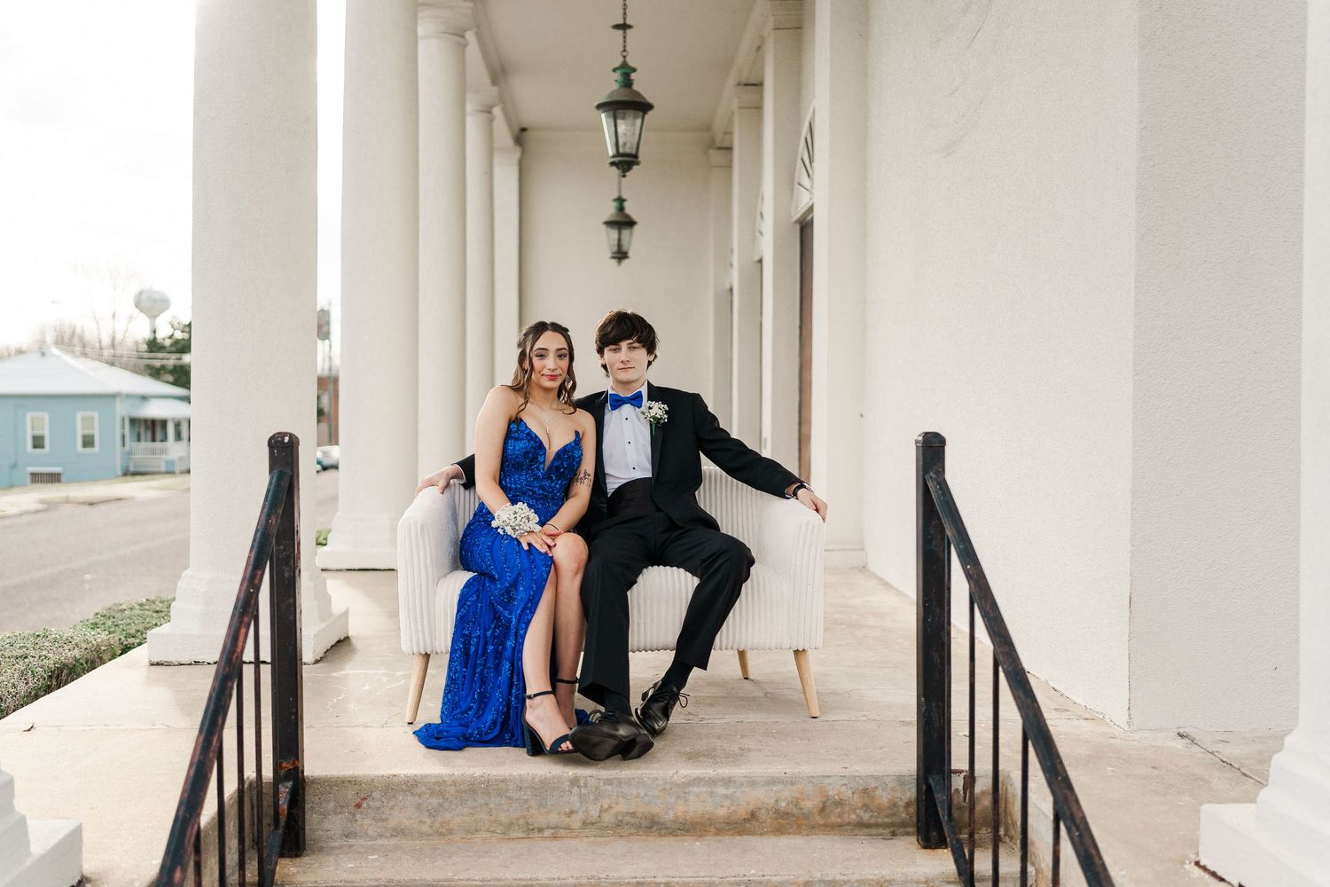 Couple sits on white porch steps in formal attire outside columned building.