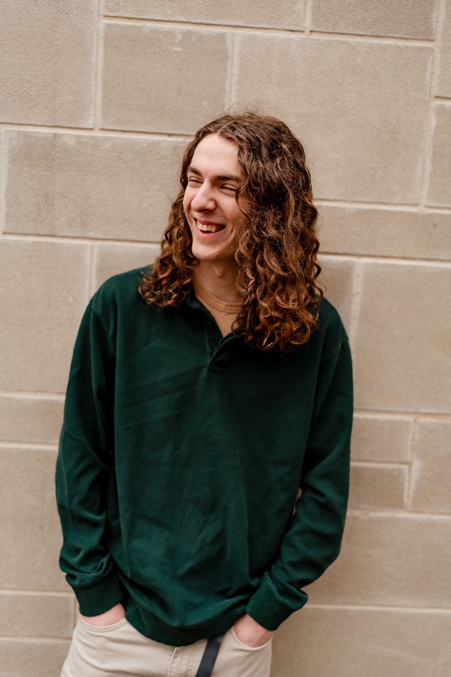 Person in forest green pullover sweater smiling against beige stone wall.