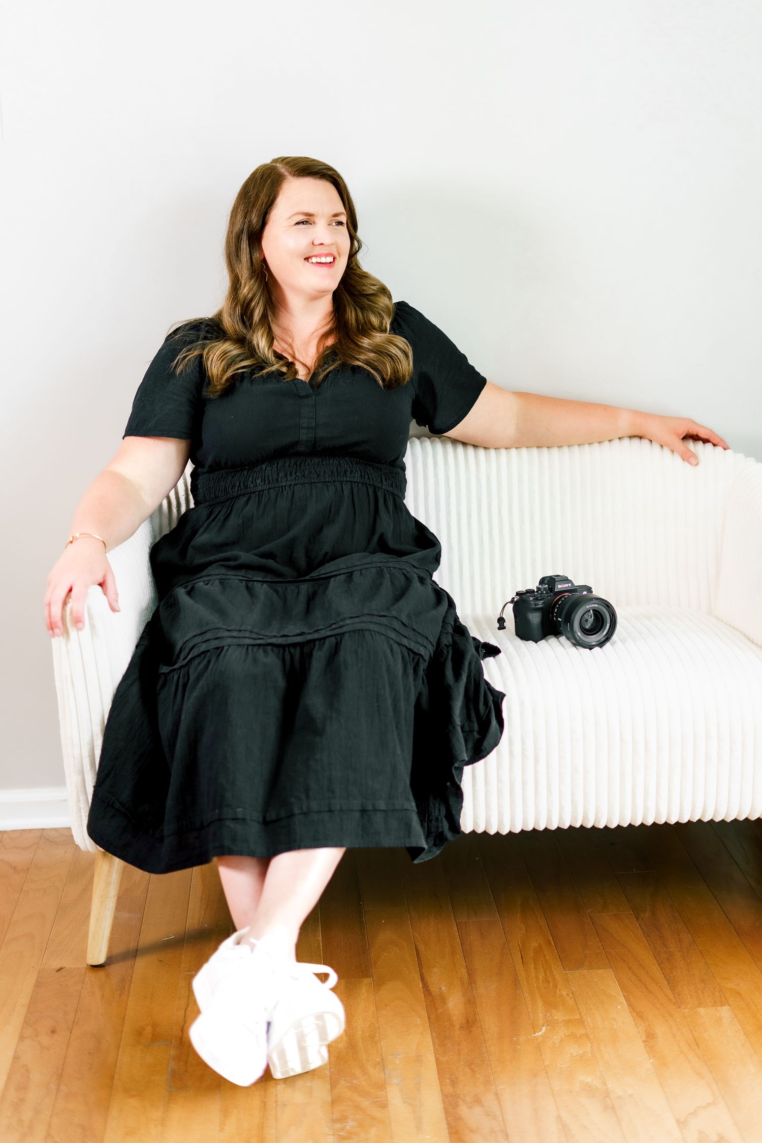 Person in flowing black dress reclines casually on white bench with camera.