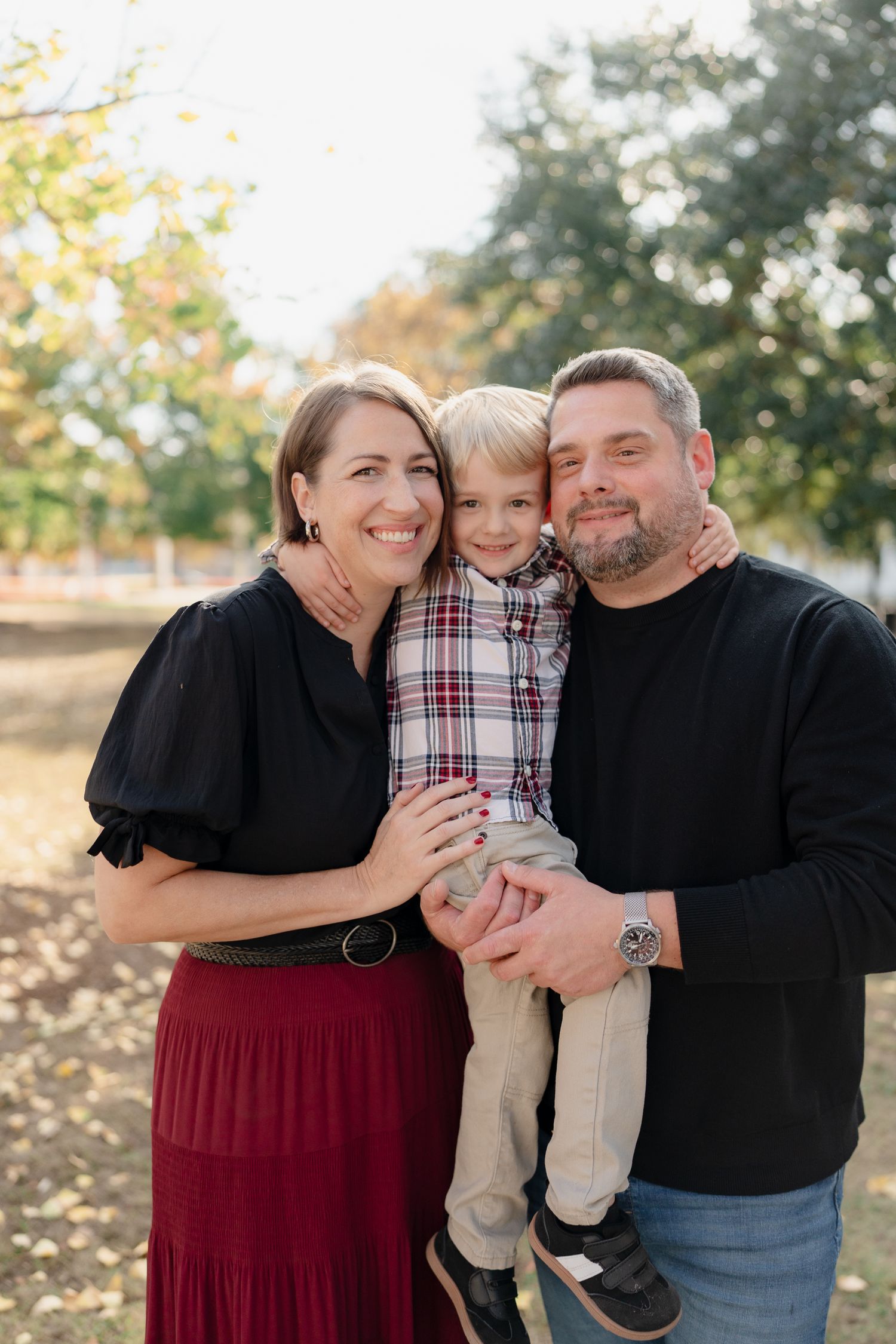 Happy family portrait with mother, father, and young son embracing outdoors in a sunny park setting.