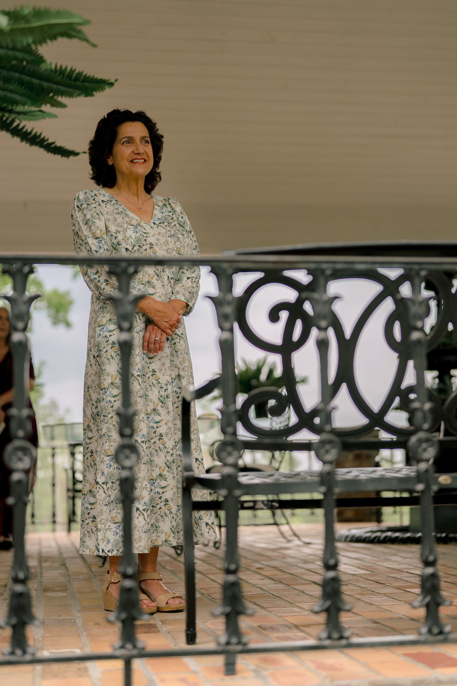 Elegant woman in floral dress standing on ornate iron balcony railing at outdoor venue.