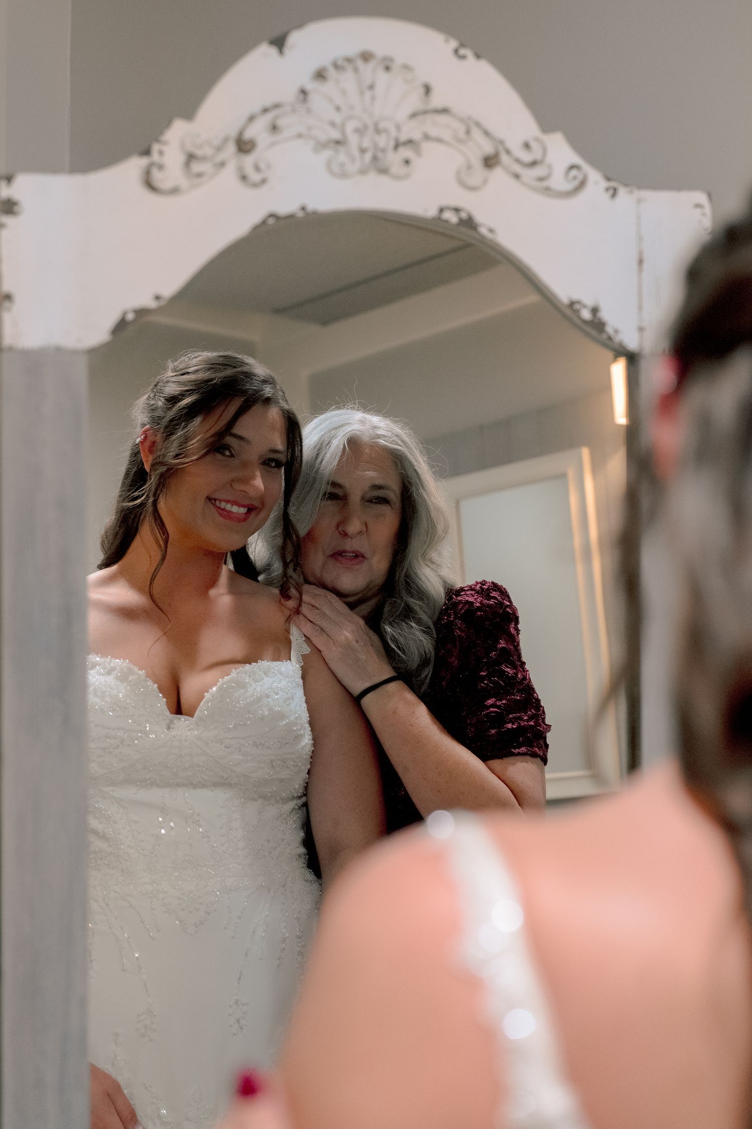 Bride in white lace gown smiles in ornate mirror as older woman adjusts her necklace during wedding preparations.
