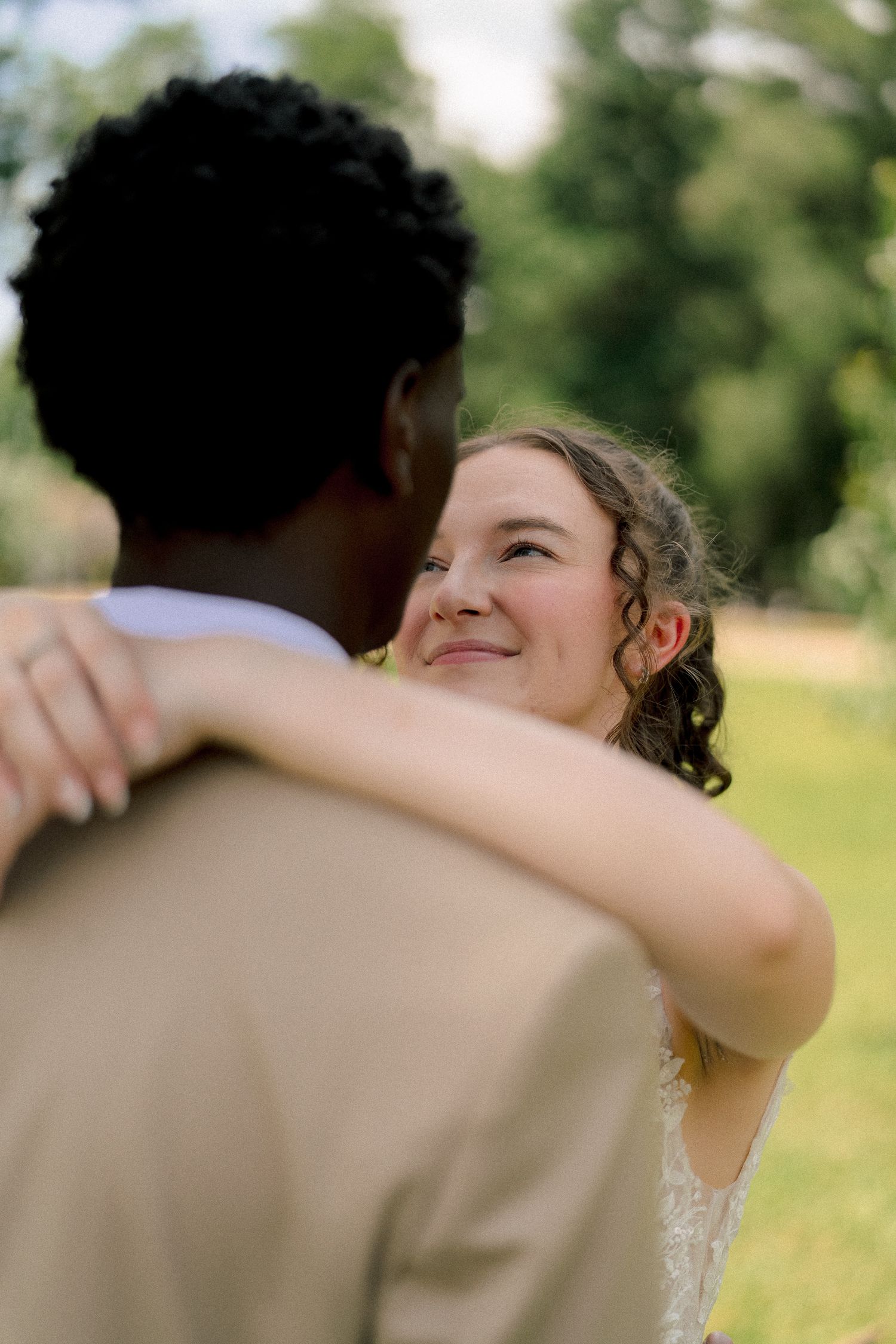 Bride in lace wedding dress smiles up at groom while embracing outdoors in lush green garden setting.