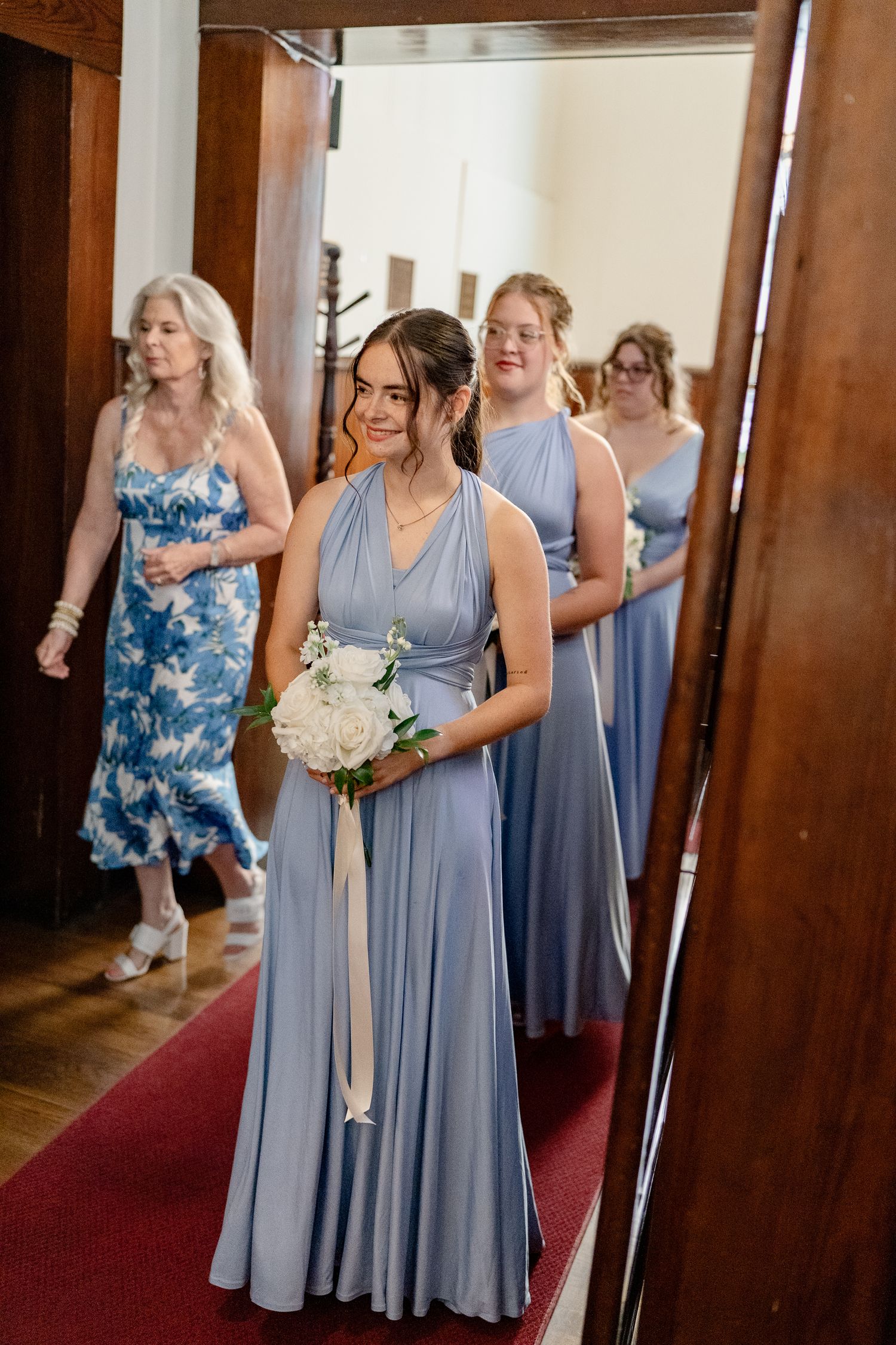 Bridesmaids in flowing periwinkle blue dresses gather in a hallway with wooden pillars and red carpet.