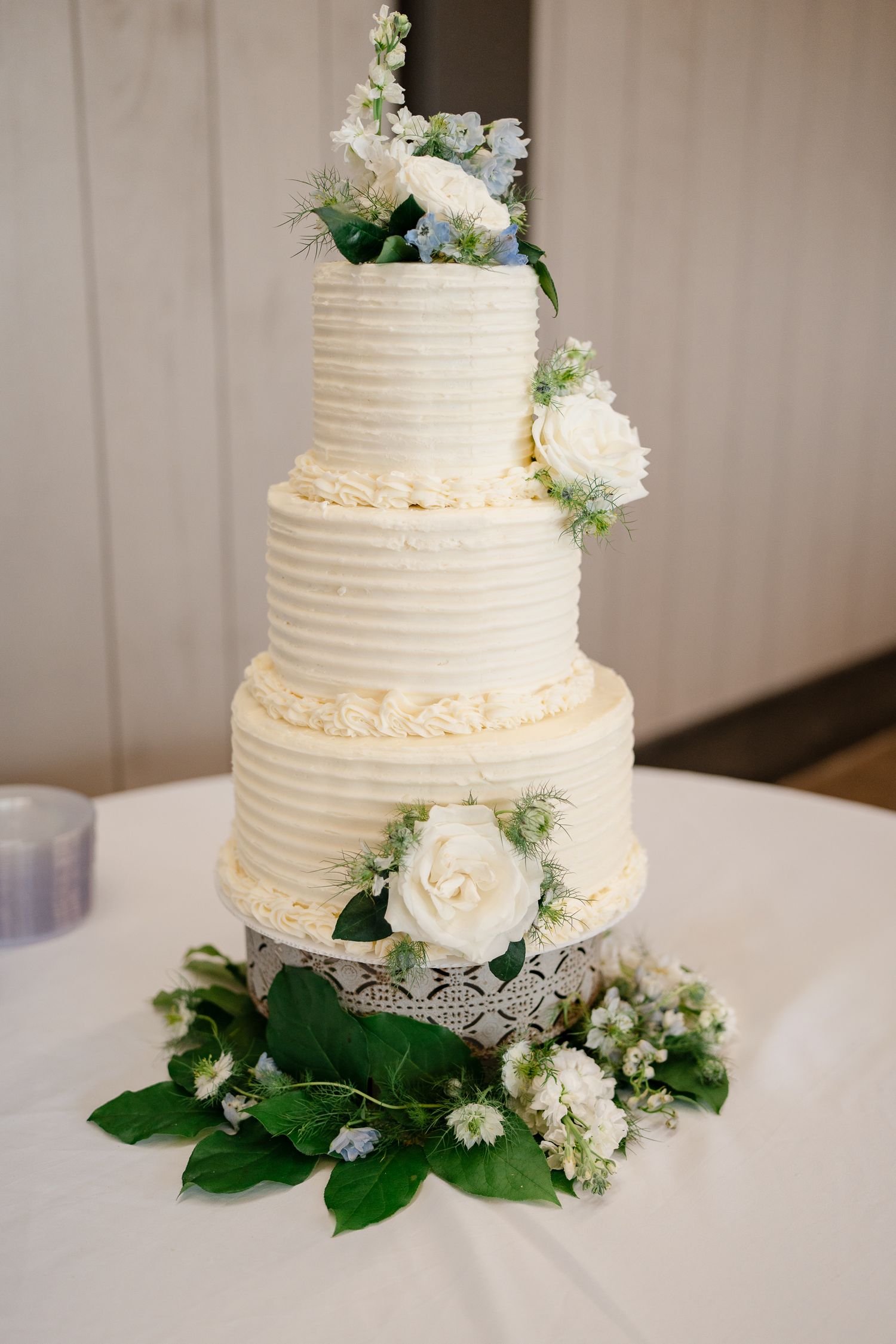 Elegant three-tiered white wedding cake decorated with fresh flowers and greenery on a white tablecloth.