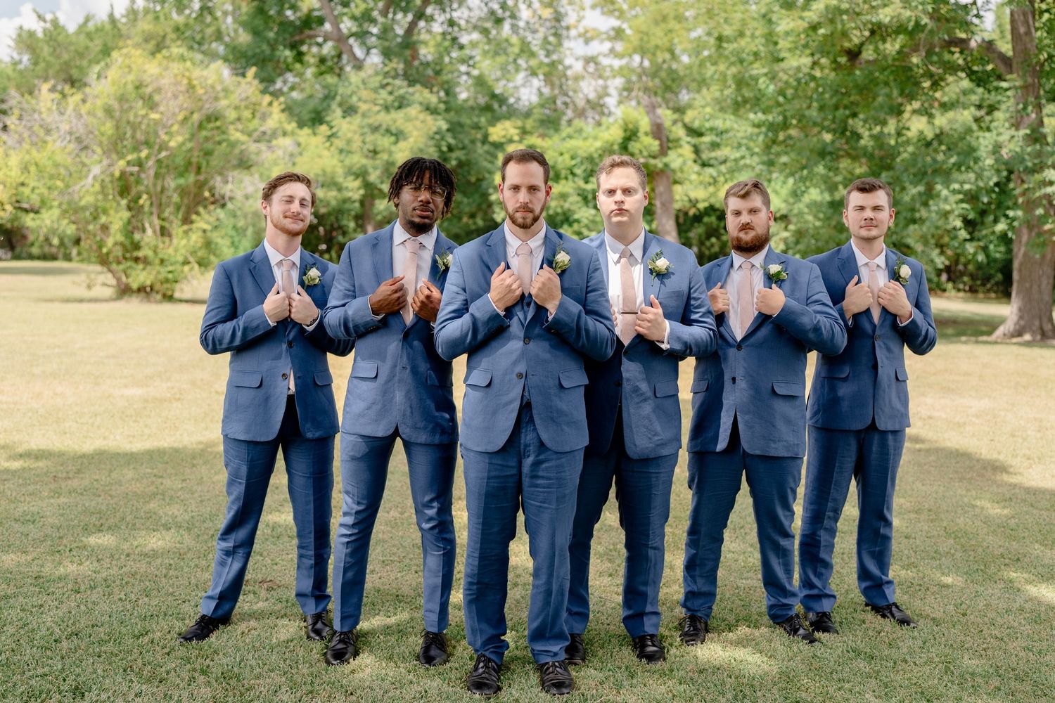 Group wearing matching navy blue suits standing together outdoors on grass with trees in background.