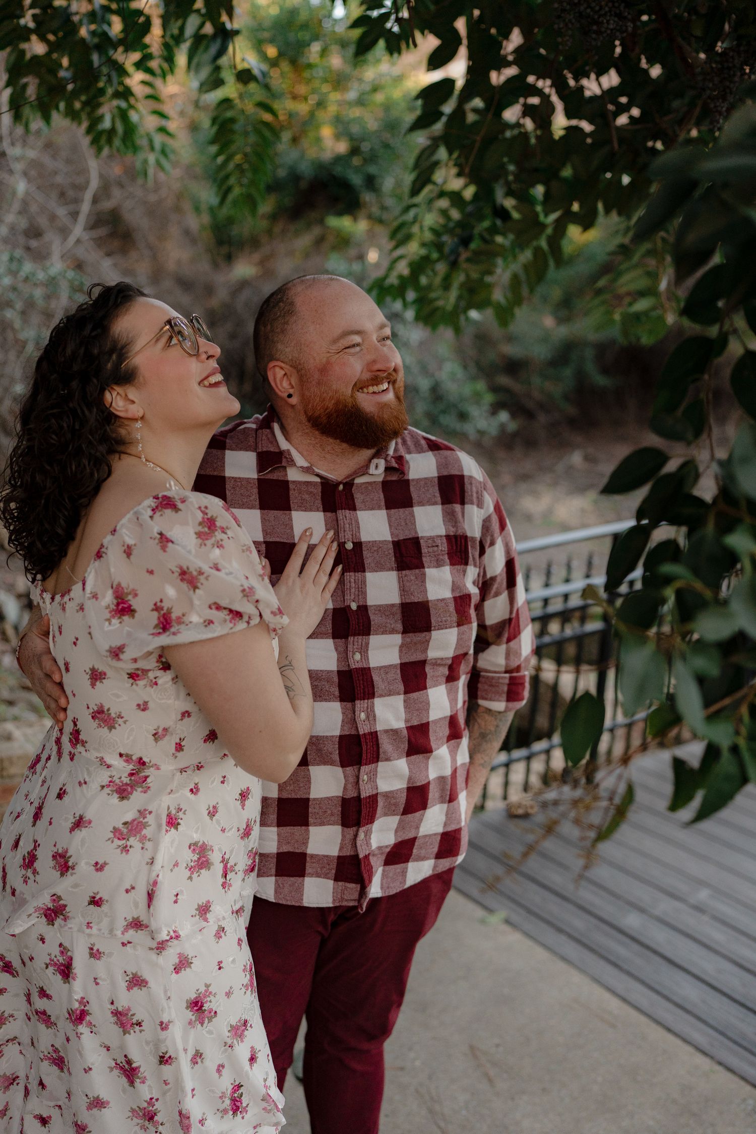 A couple wearing burgundy plaid and floral prints share joyful laughter while standing outdoors under trees.