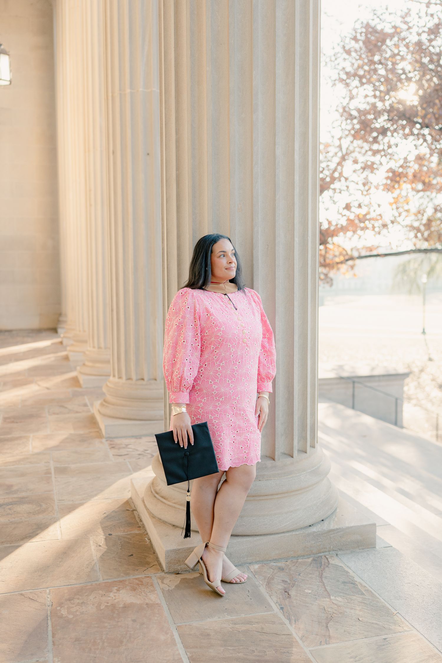 Graduate in pink dress poses between classic white columns in elegant architectural setting.