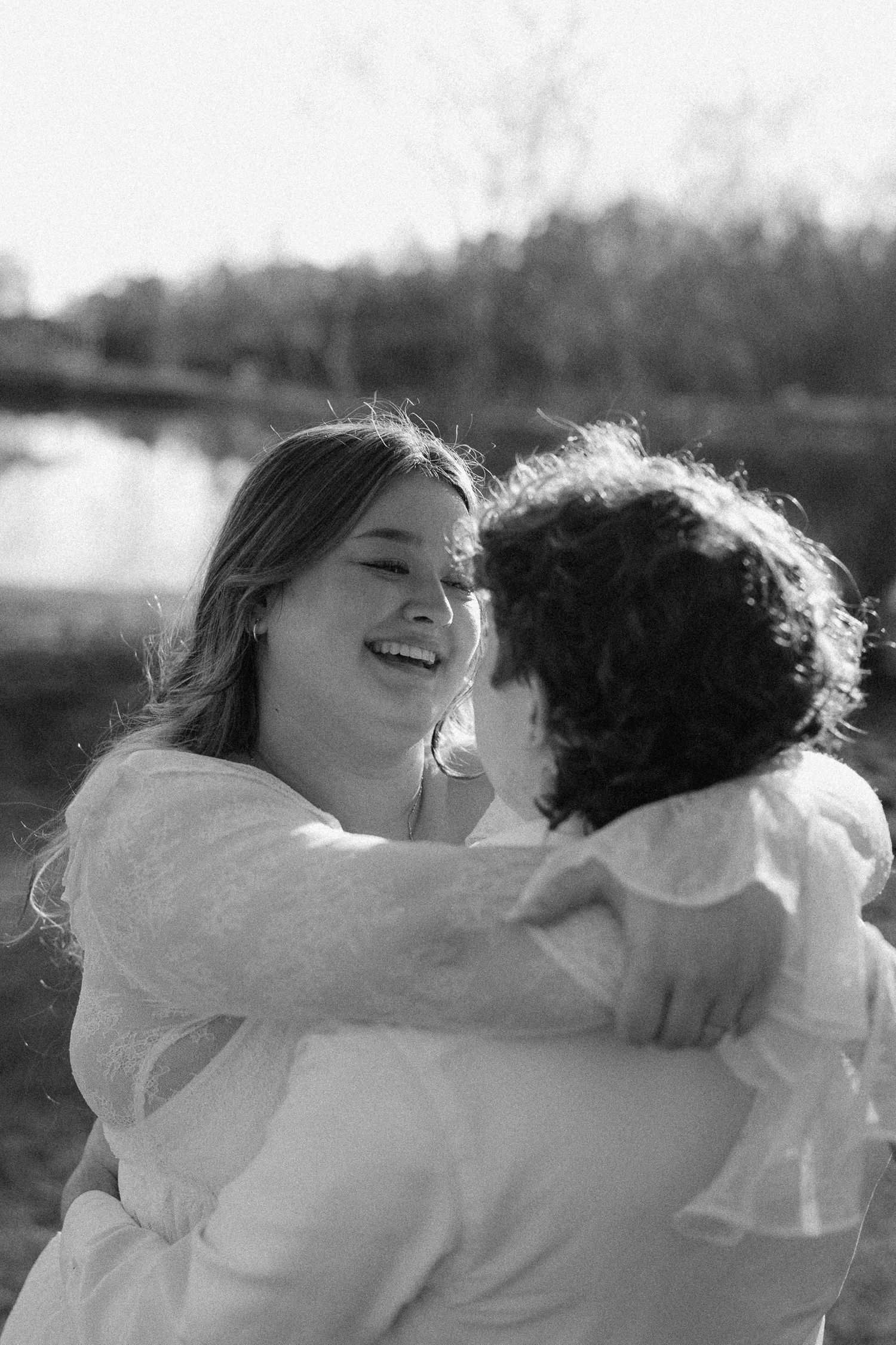 Black and white image of laughing woman embracing her partner near a reflective lake.