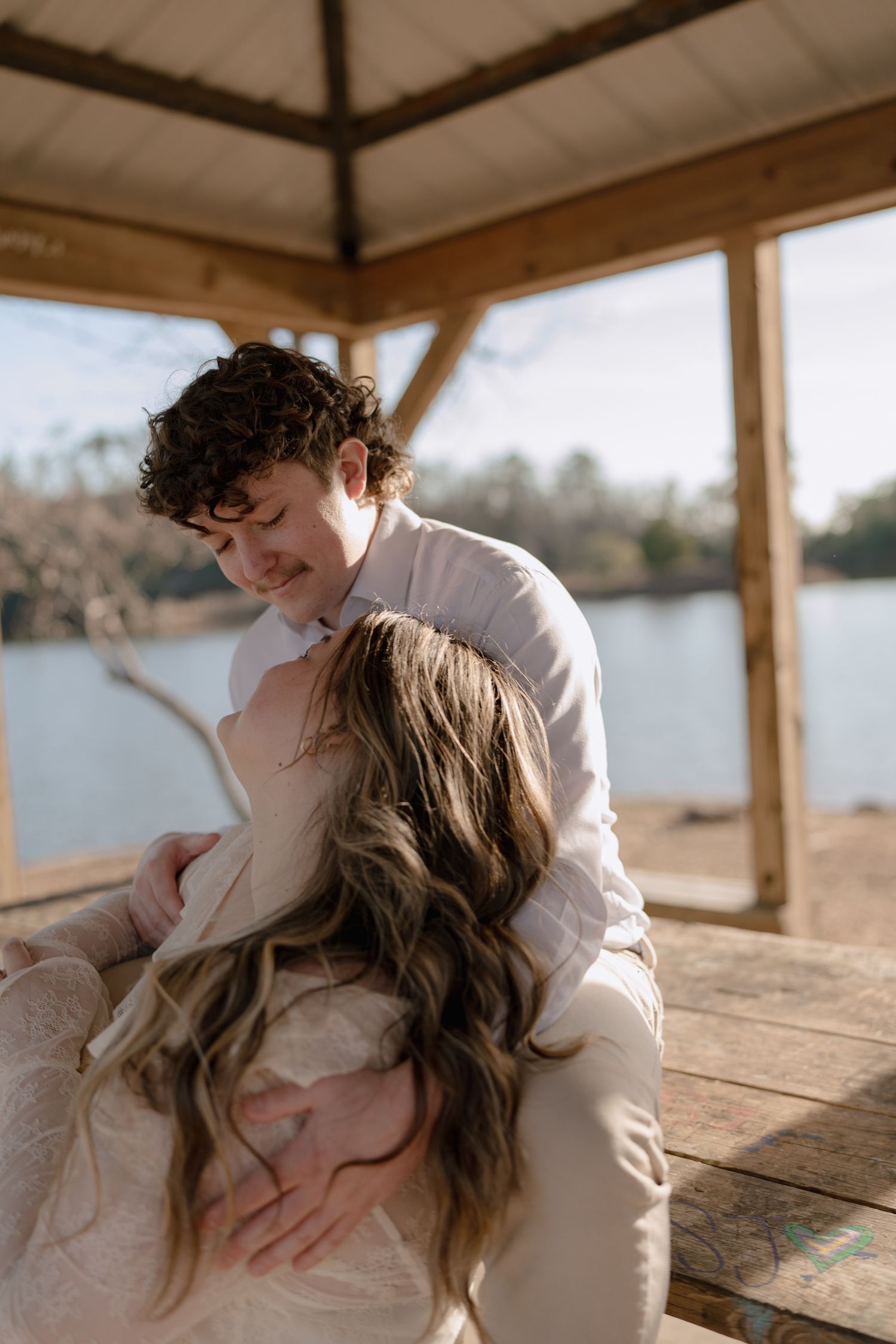 Couple sharing a tender embrace under a rustic wooden gazebo overlooking a calm lake.