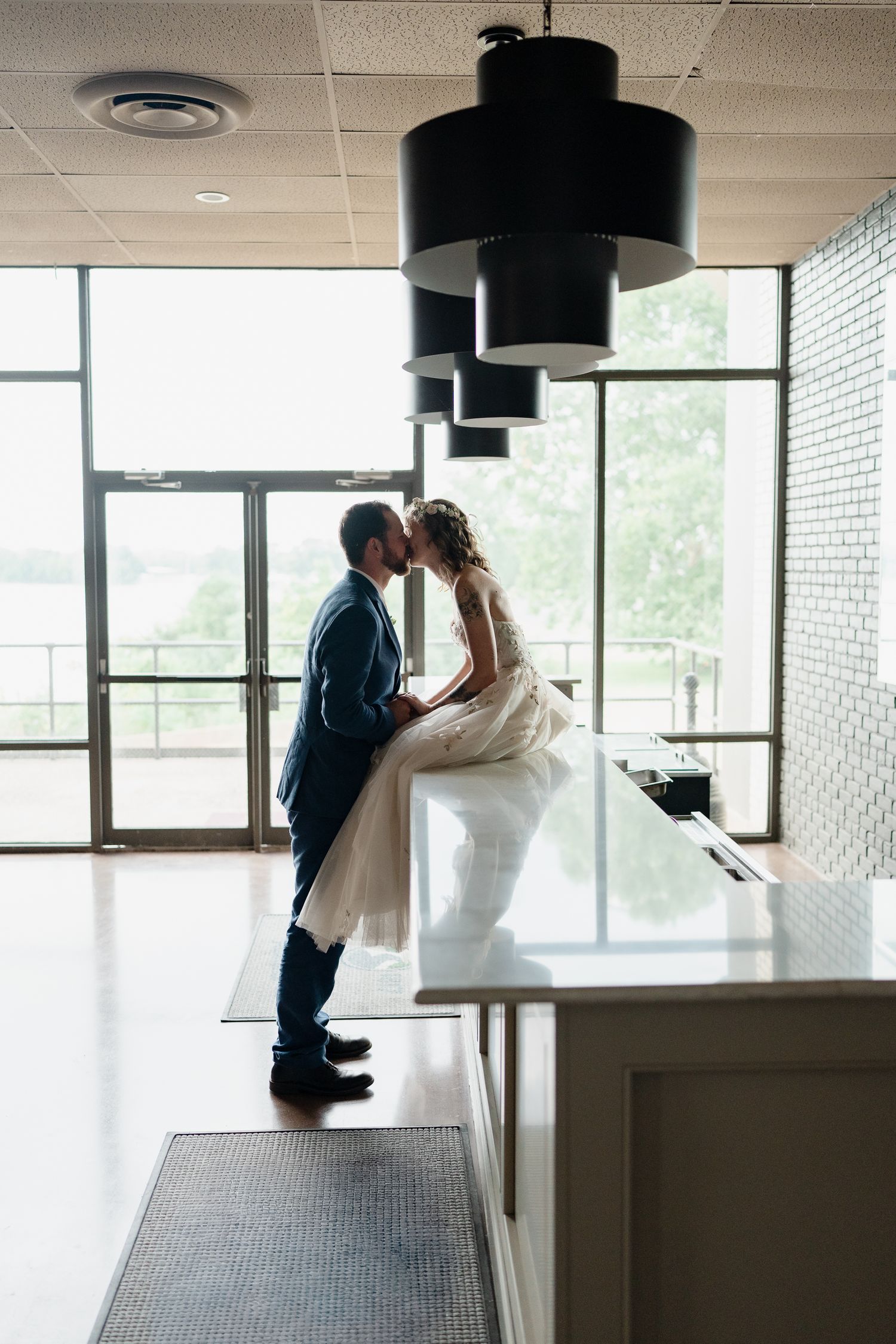 A romantic couple shares an intimate moment in a modern office building hallway with large windows and pendant lights.