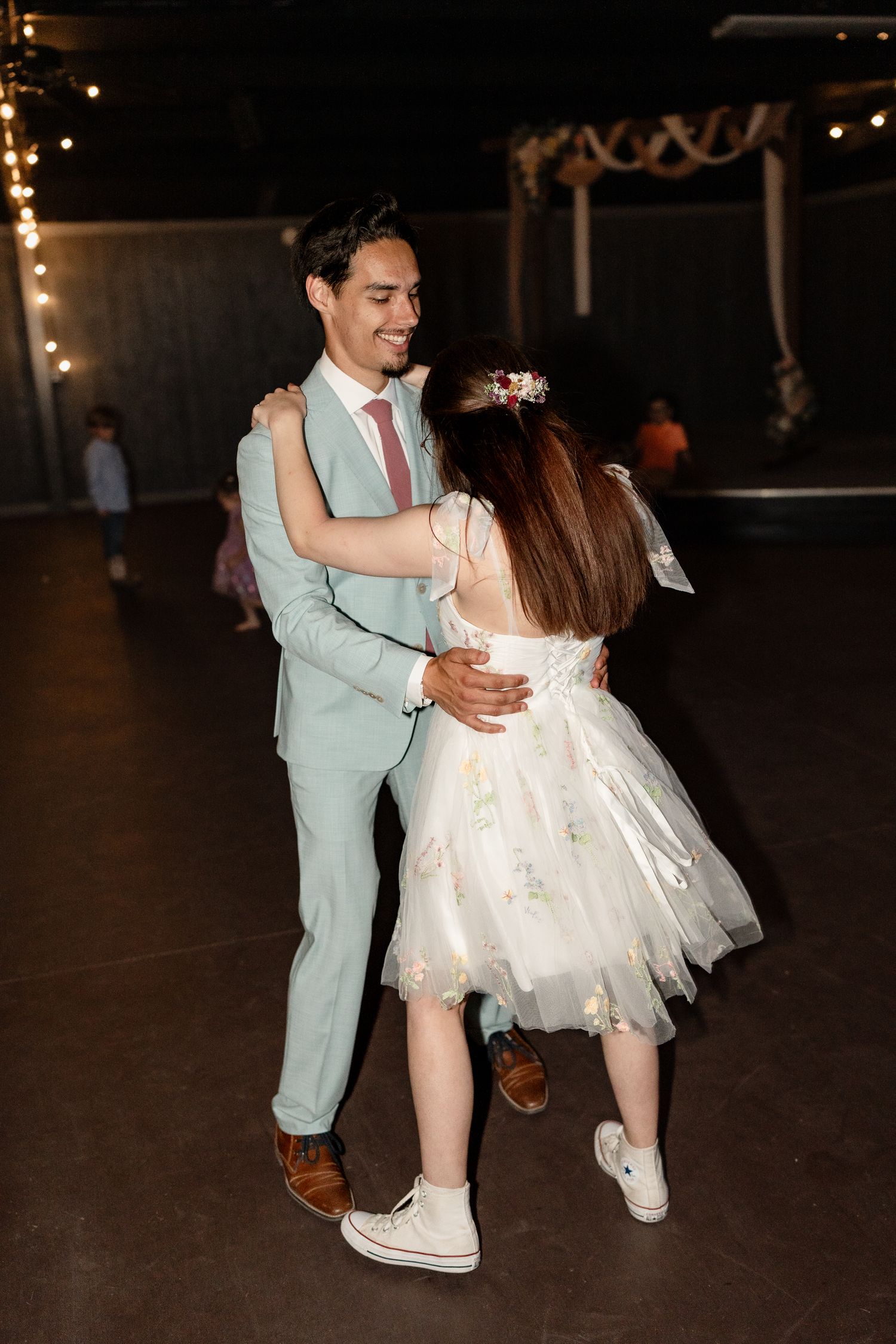 A couple dances together at a wedding reception, with the bride wearing a short white dress and the groom in a light blue suit.