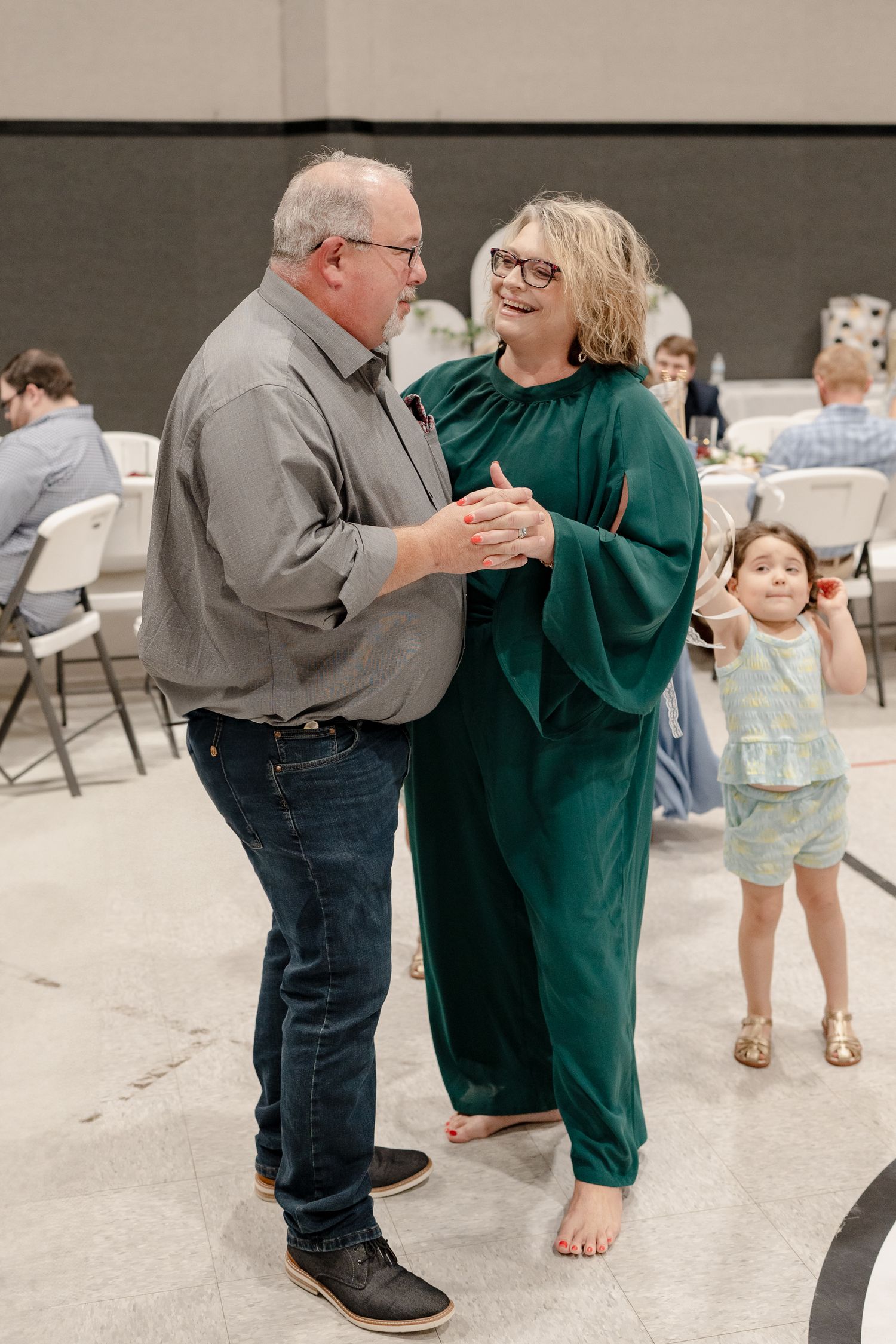 People dancing together in a community hall with folding chairs visible in the background.