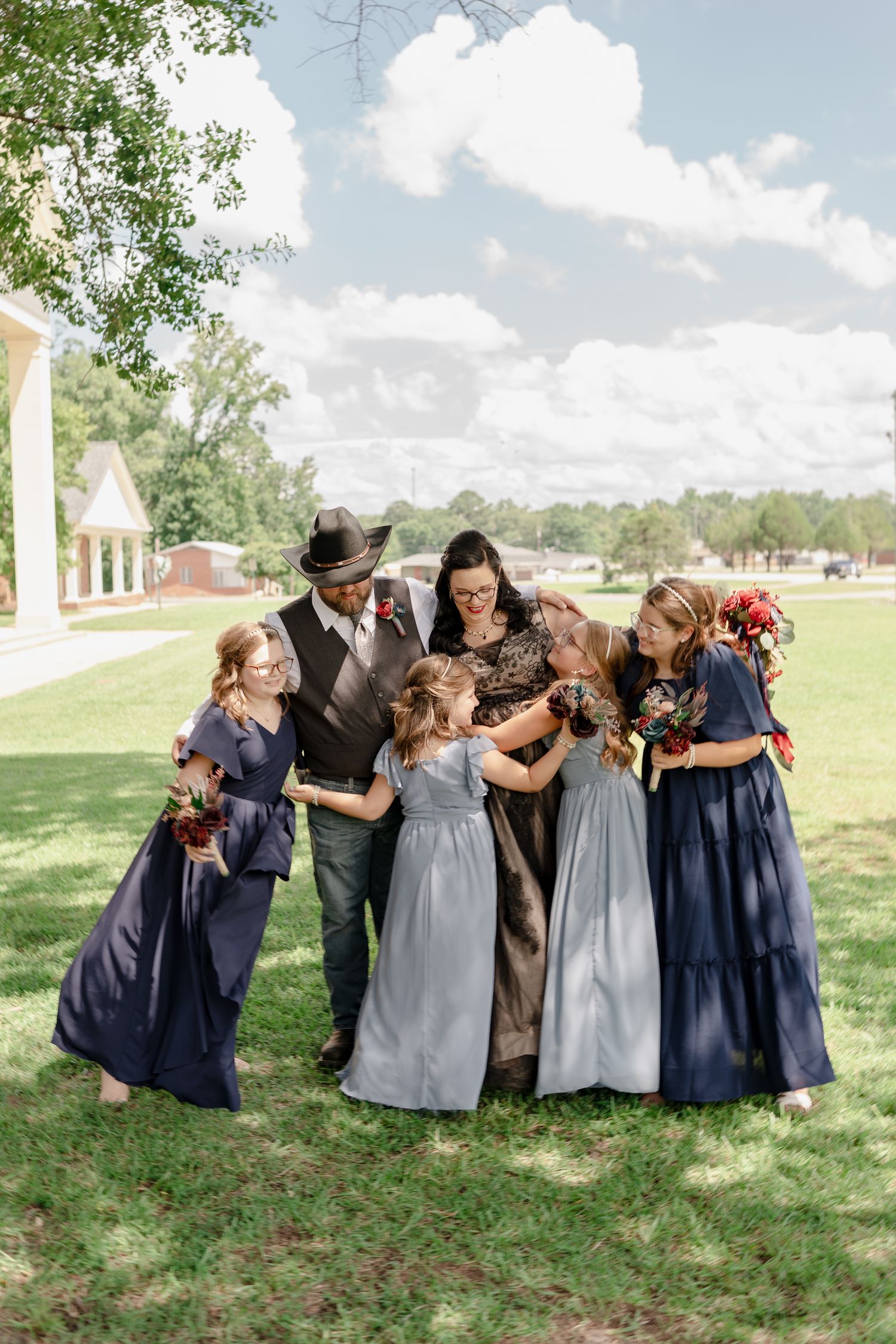 Candid moment of a wedding party in navy and gray dresses sharing laughter on a sunny day outdoors.