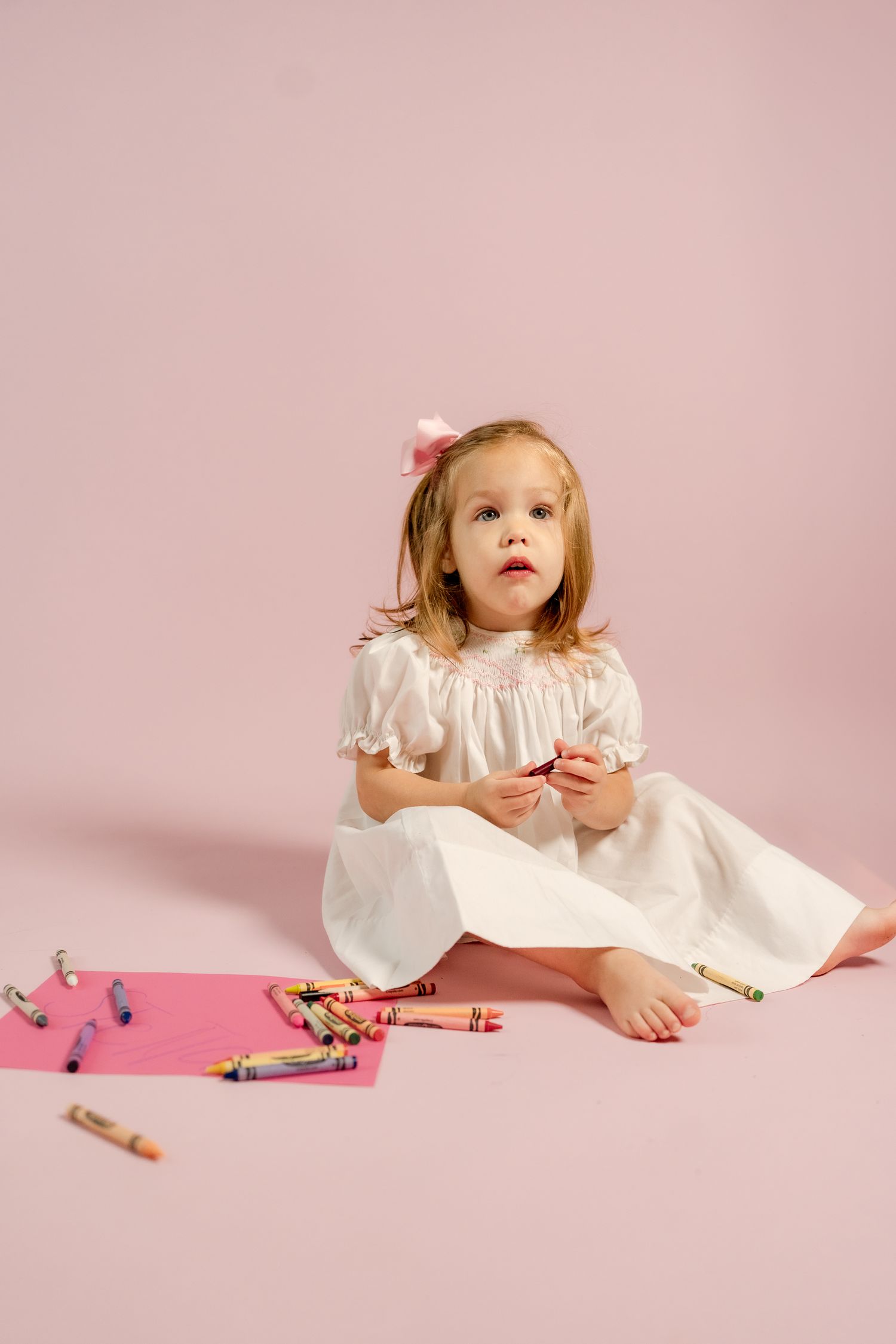 A toddler in a white dress sits playfully on a pink background with scattered crayons.