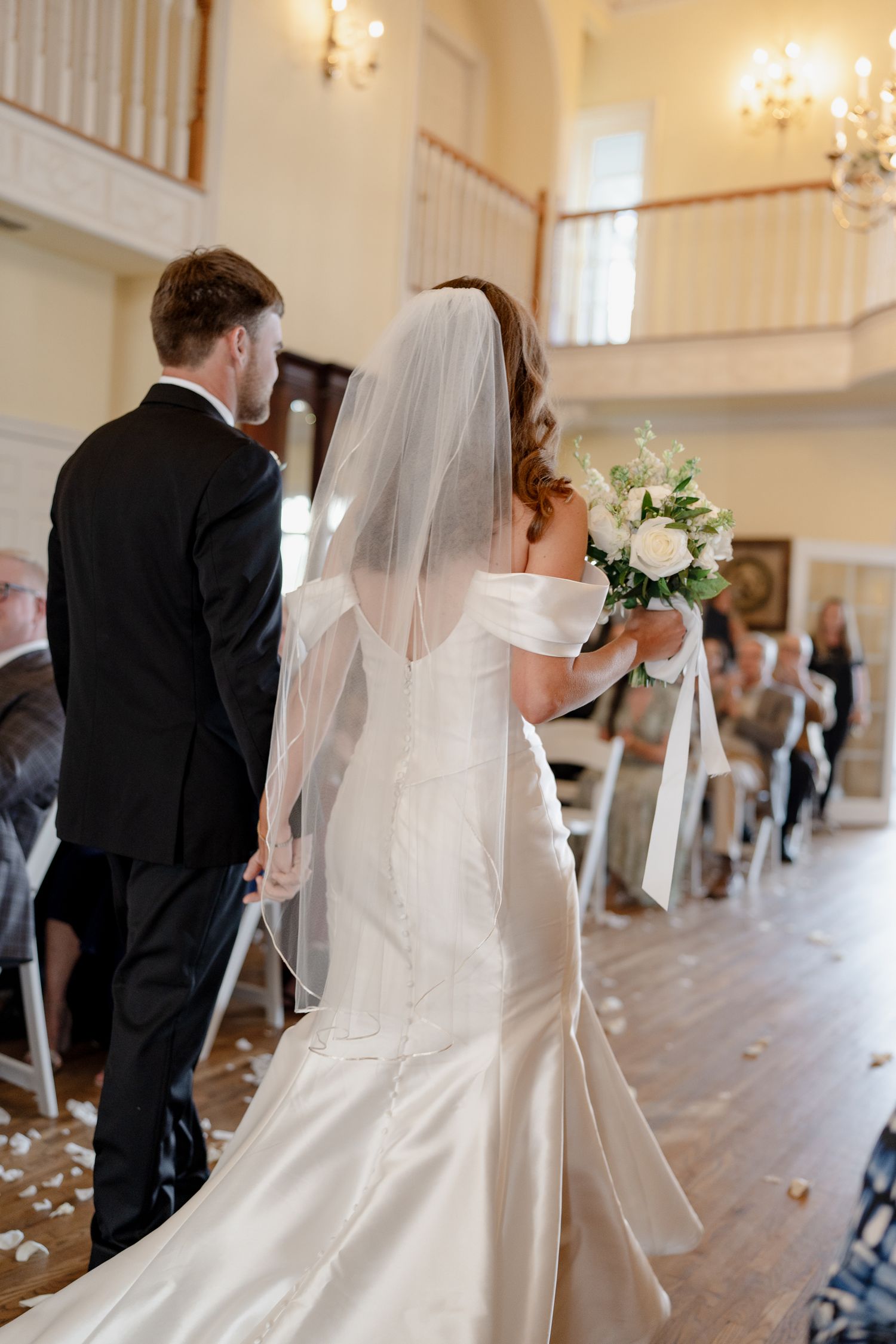 Couple walks down aisle in elegant venue with cascading white wedding gown and classic black tuxedo.